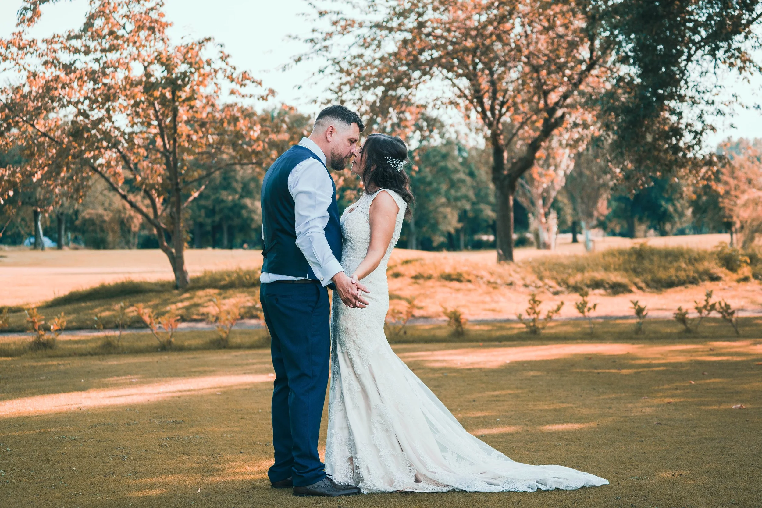 A bride and groom holding hands and about to kiss outdoors on a grassy area with trees and autumn foliage in the background.