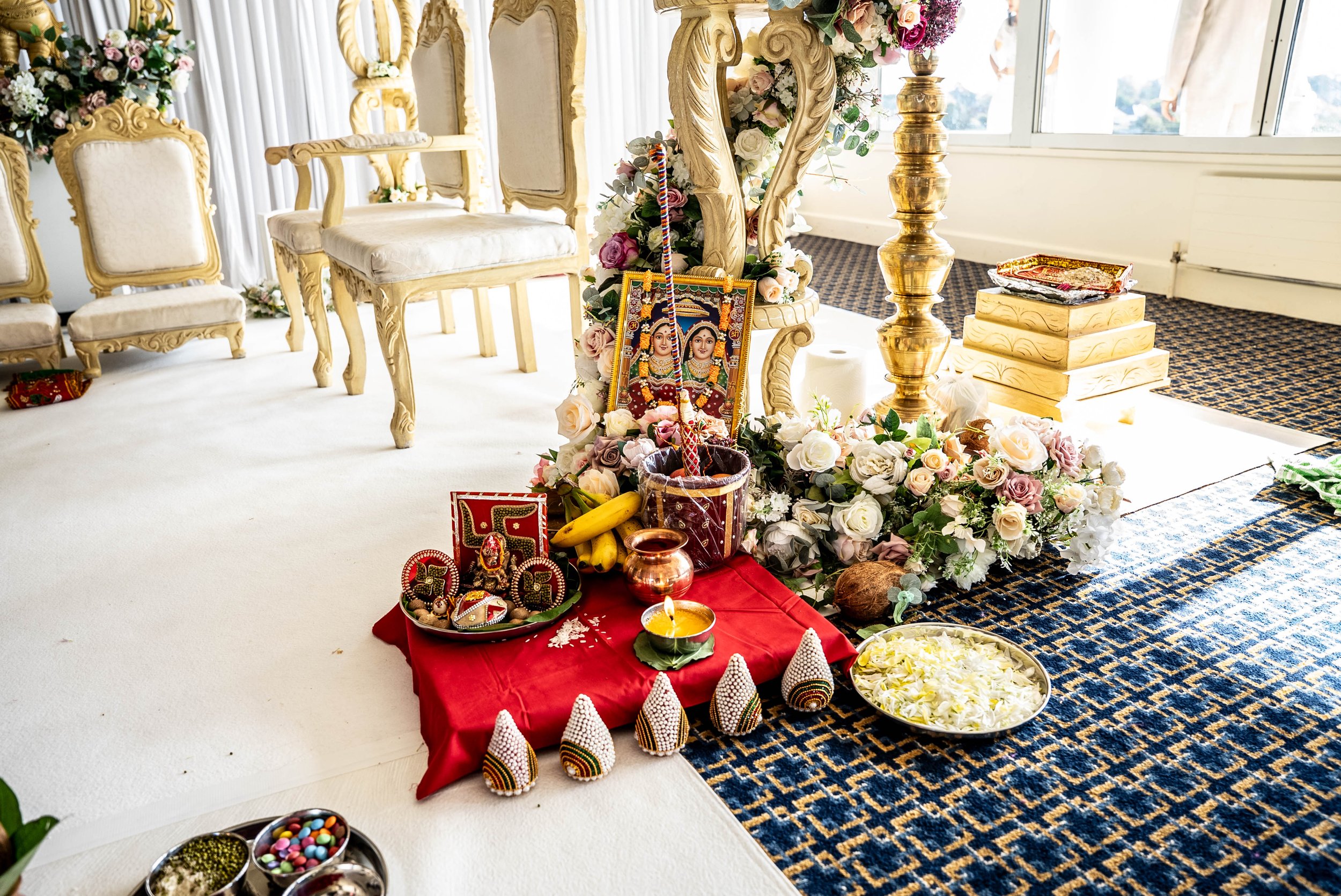 Hindu prayer setup with flowers, images of deities, and offerings on a red cloth in a well-lit room with chairs and windows.