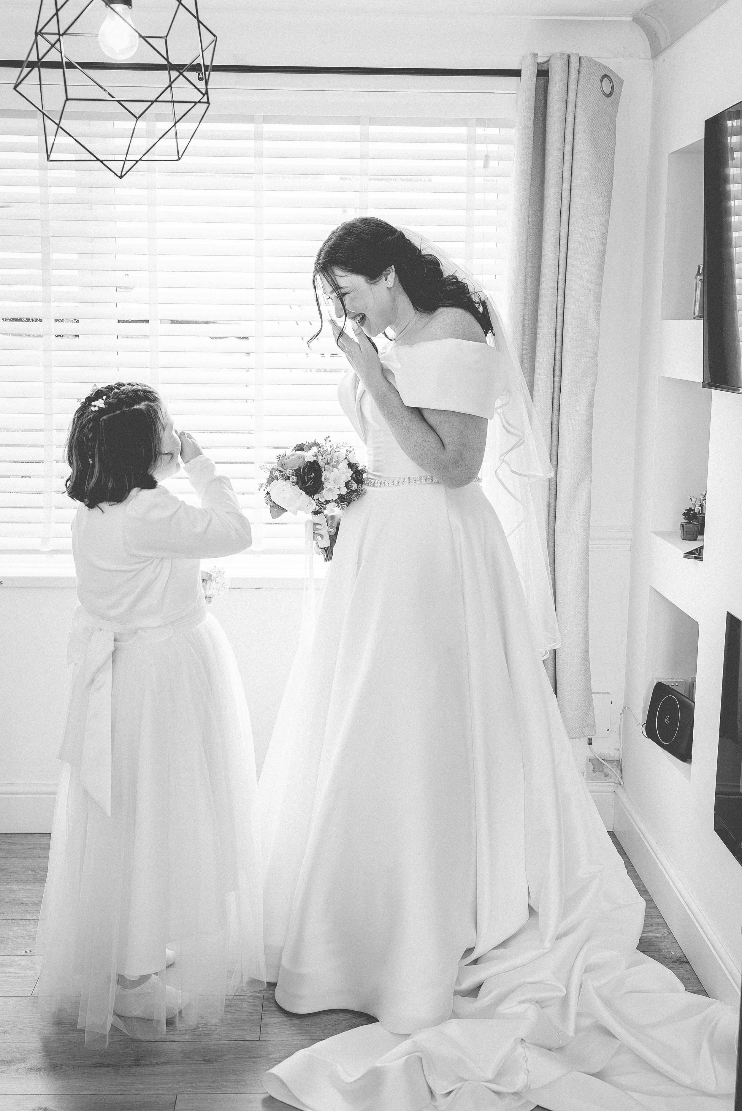 A smiling woman in a wedding dress holding a bouquet, standing in a room with a young girl offering her a flower. Both are indoors with sunlight coming through the window blinds.