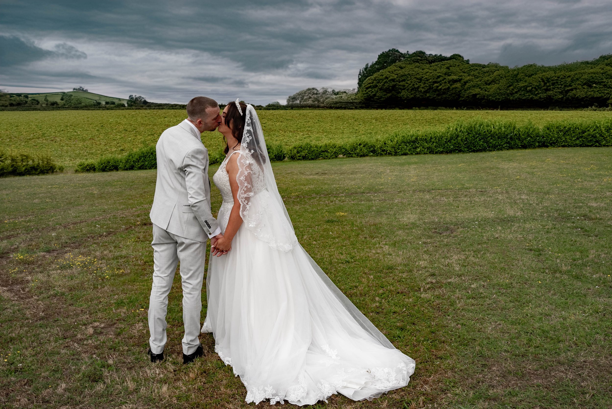 A bride and groom kissing outdoors on a grassy field with a cloudy sky and trees in the background.