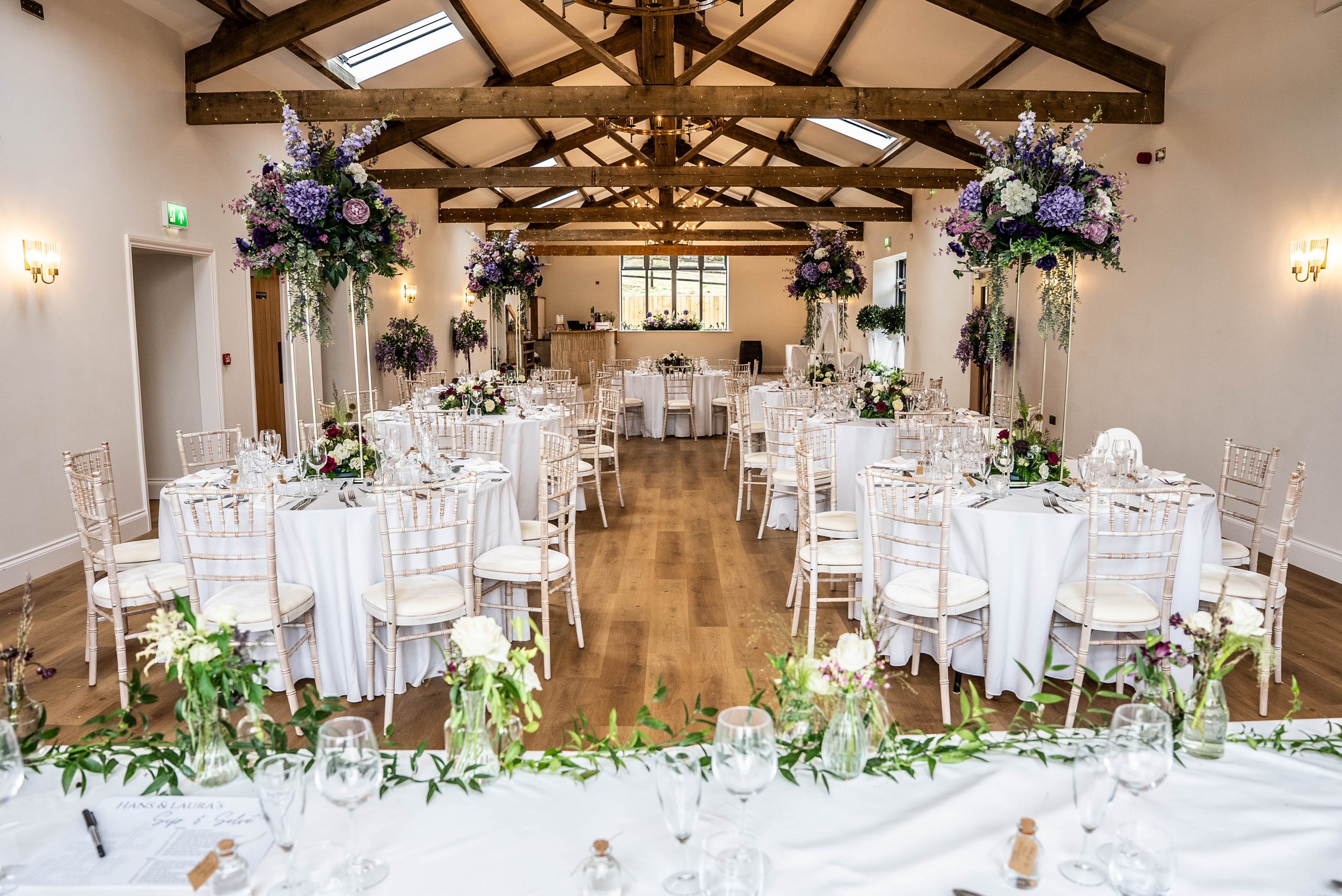 Elegant wedding reception with round tables draped in white cloth, decorated with floral centerpieces, set with glassware, and surrounded by white chairs in a spacious room with wooden beams and skylights.