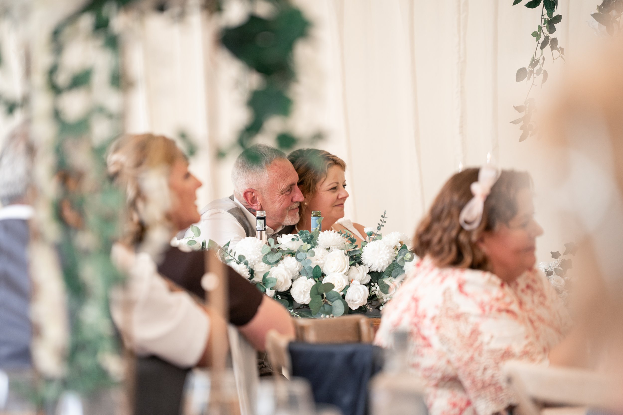 People attending a formal event or celebration, sitting at a table decorated with white flowers and greenery, with a light-colored wall and hanging greenery in the background.