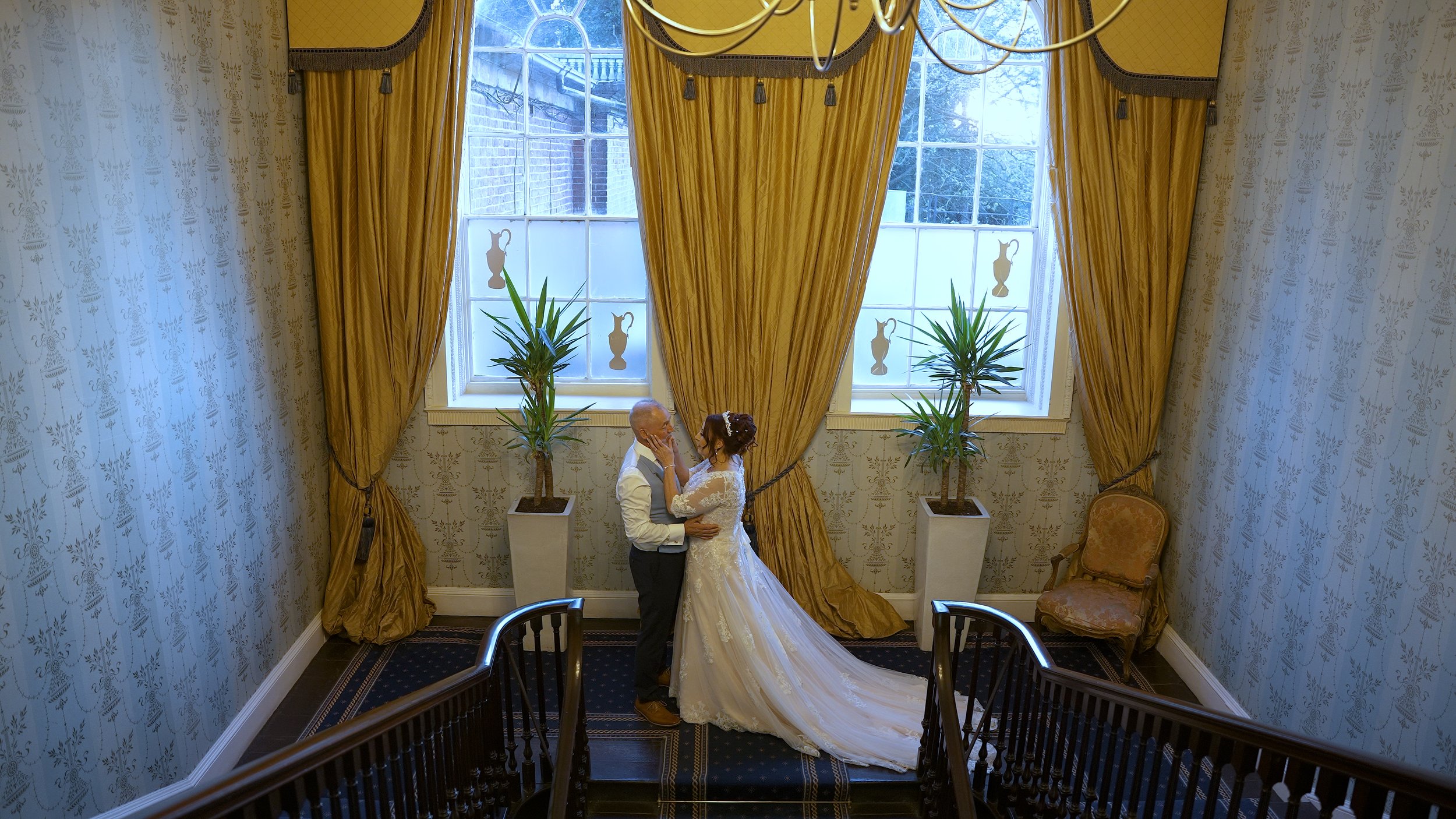 A bride and groom stand close together in a decorated room with gold curtains and potted plants, sharing an intimate moment.