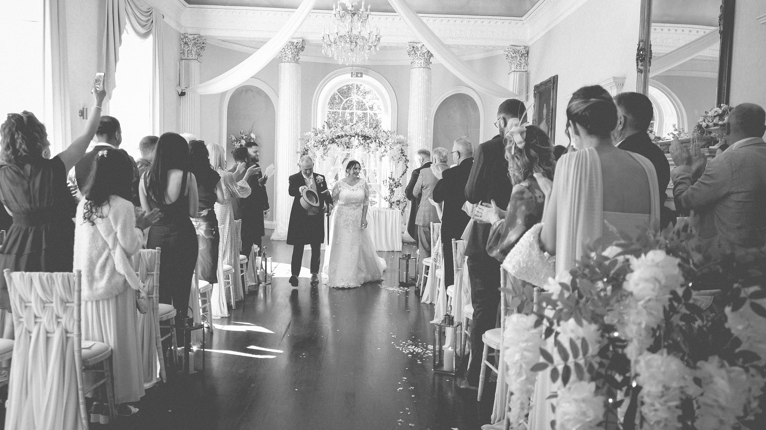 Black and white photo of a wedding ceremony inside an elegant, decorated hall with tall windows, draped fabric, and a chandelier. The bride and groom are walking down the aisle, surrounded by guests.