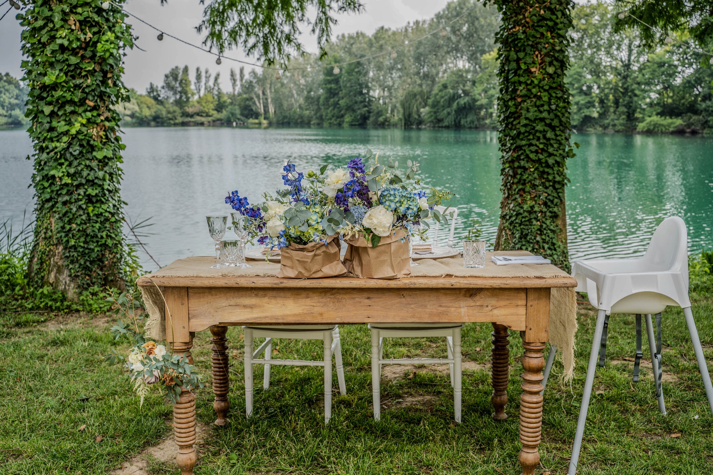 A wooden outdoor dining table decorated with a blue and white floral centerpiece, set with glassware, on a grassy area by a lake with trees, hanging string lights, and two large tree trunks covered in ivy.