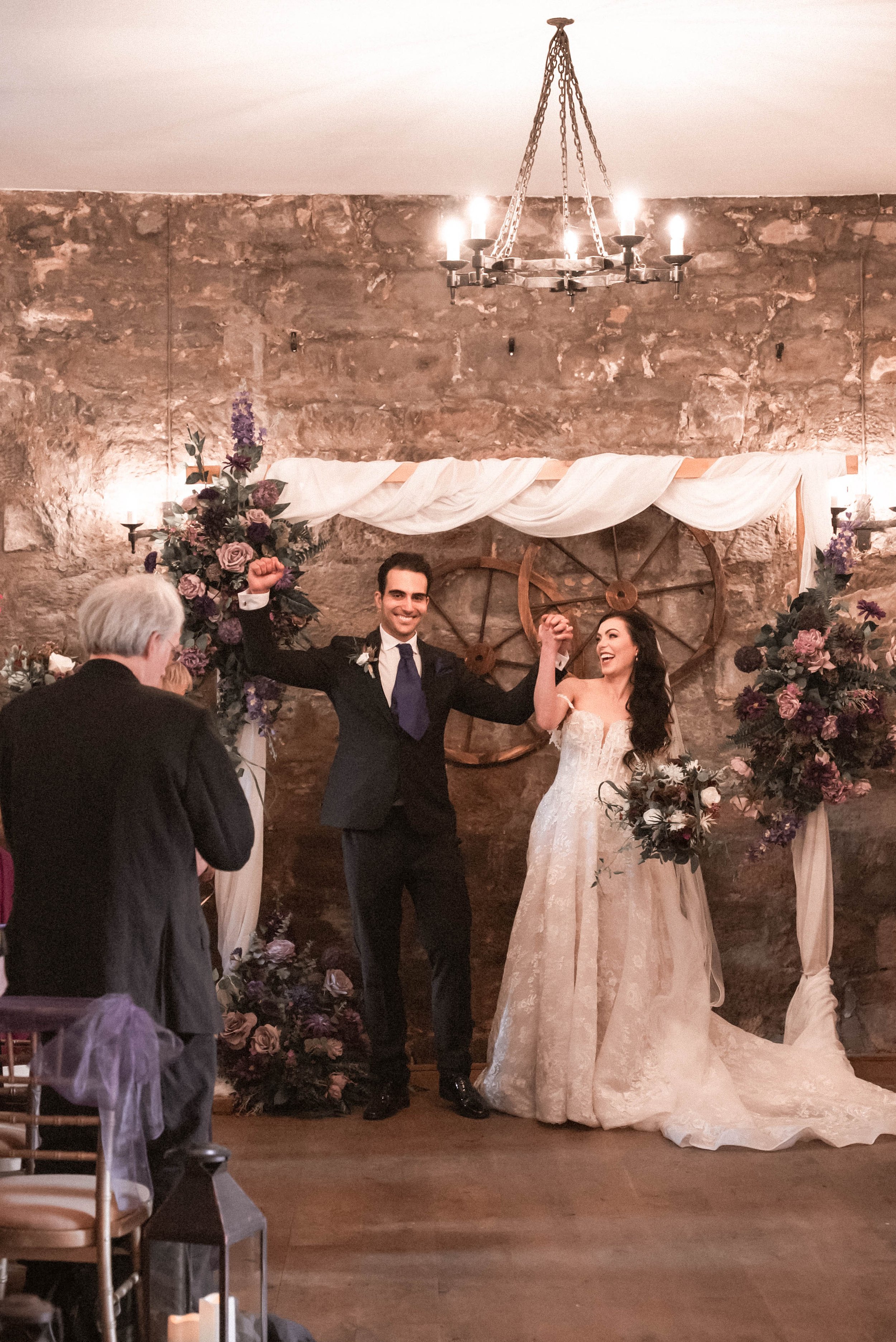 Bride and groom celebrating their wedding, holding hands up, in front of a rustic stone wall decorated with flowers and wagon wheels.