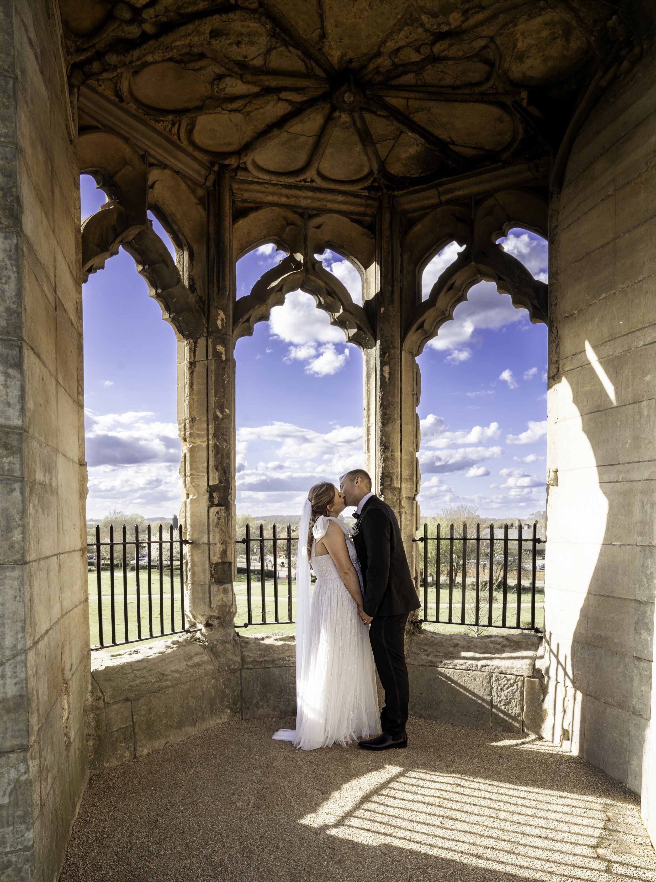 A bride and groom sharing a kiss inside a stone archway with Gothic architectural details, overlooking a sunny landscape with blue sky and white clouds.