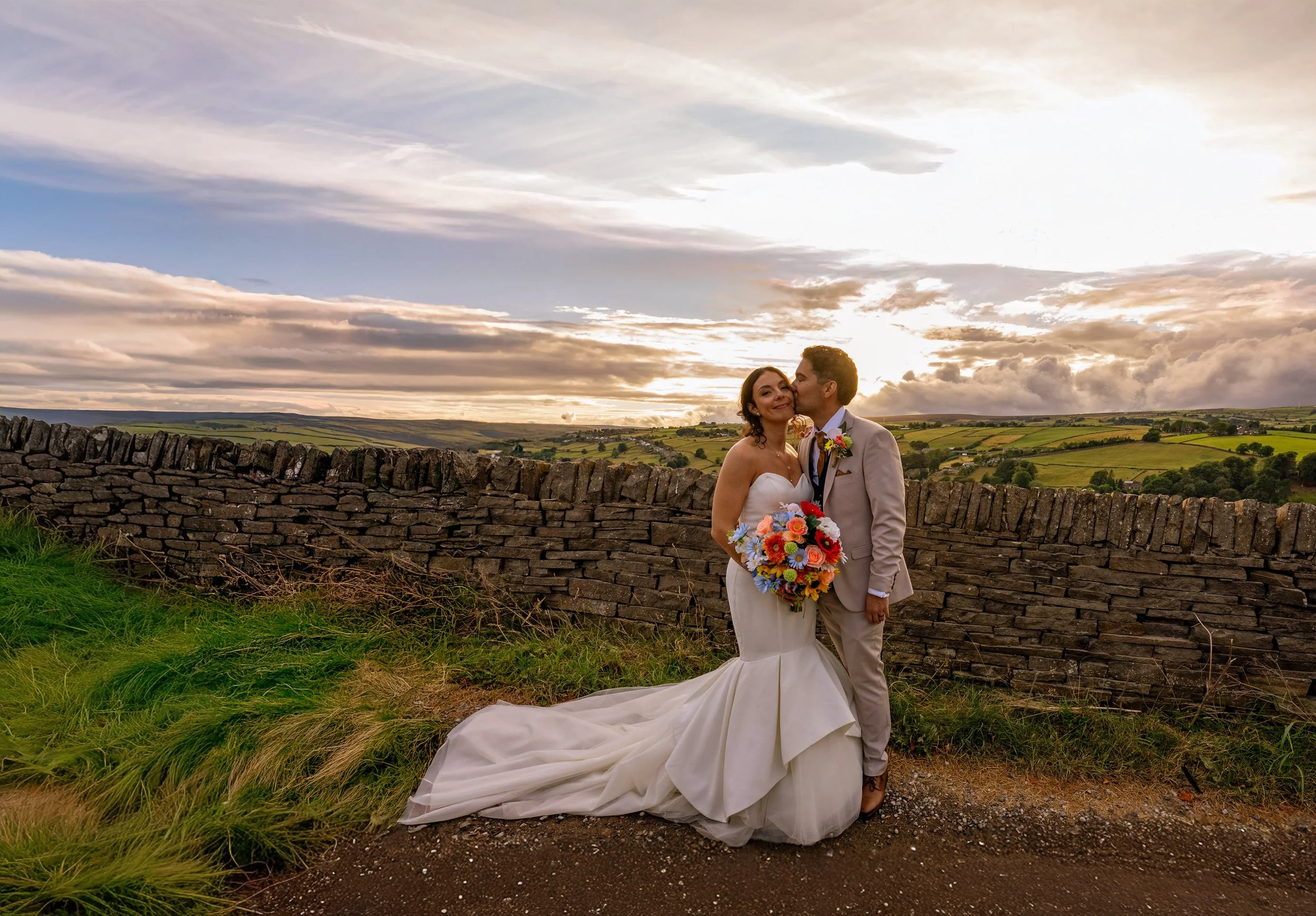 A bride and groom standing outside near a stone wall, with green fields and a sunset sky in the background, sharing a tender moment.