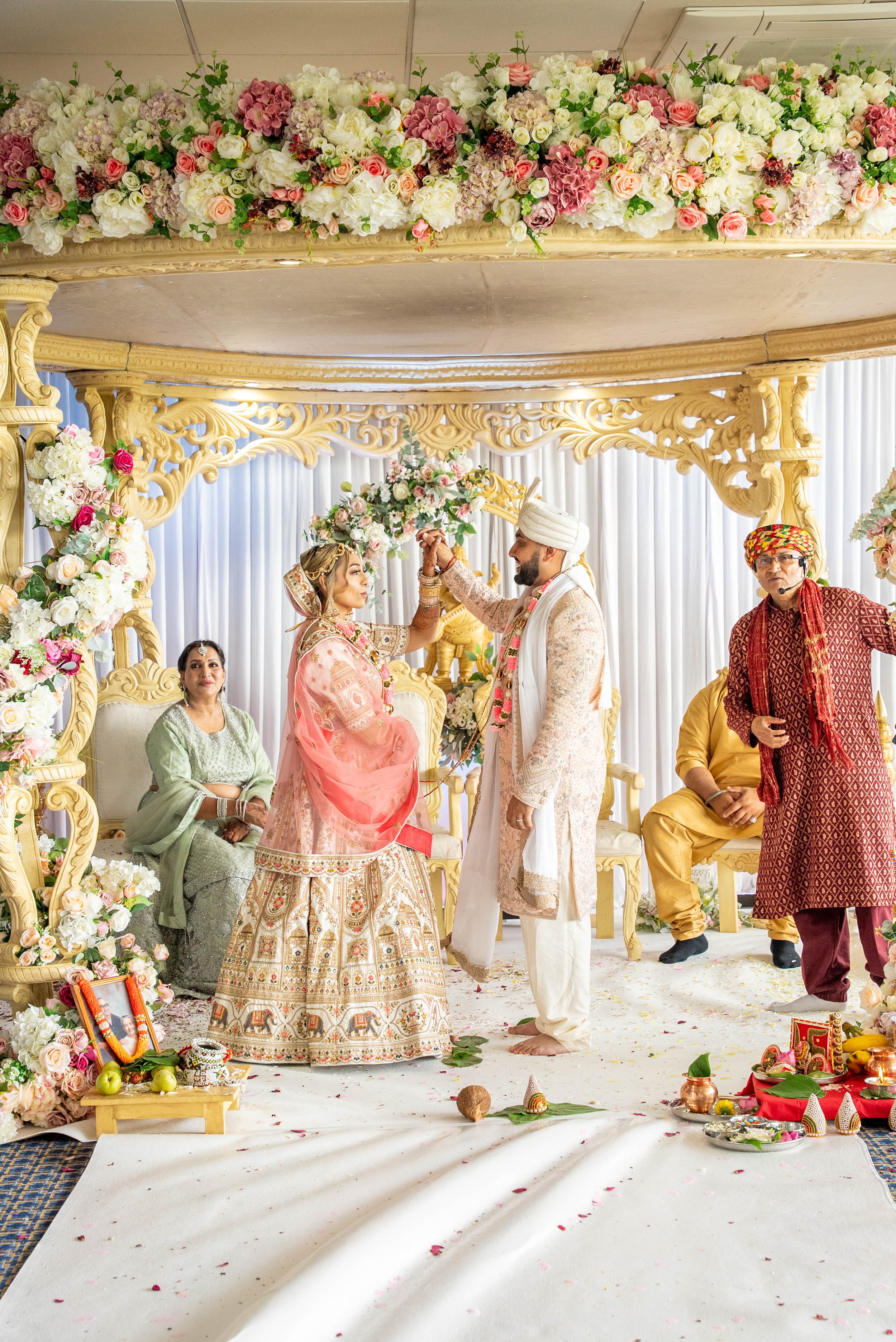 A traditional Indian wedding ceremony with bride and groom exchanging garlands, surrounded by floral decorations, family members, and ceremonial items.