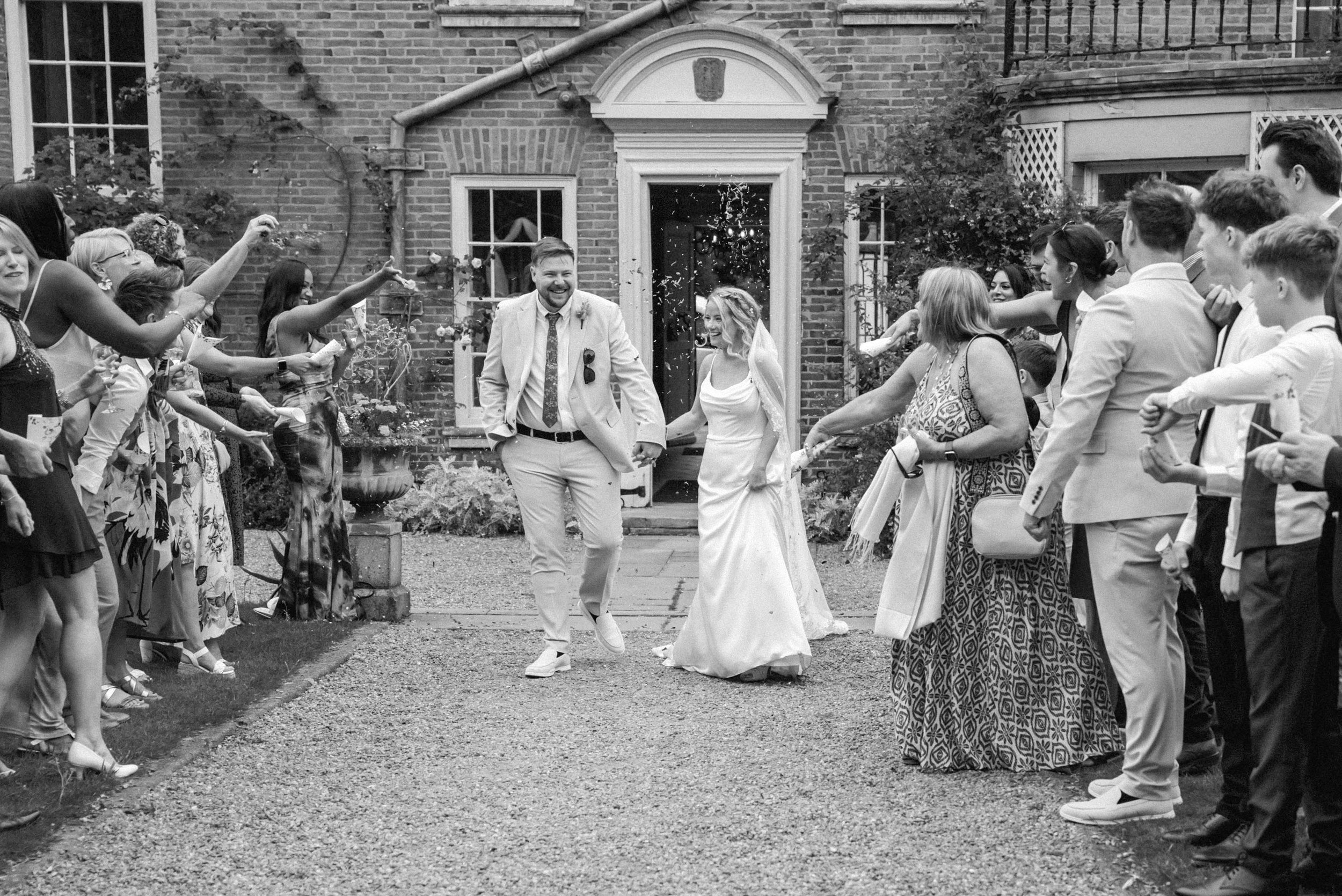 A black and white photo of a wedding celebration showing a bride and groom walking hand in hand through a crowd of guests who are throwing confetti and celebrating outside a brick building.