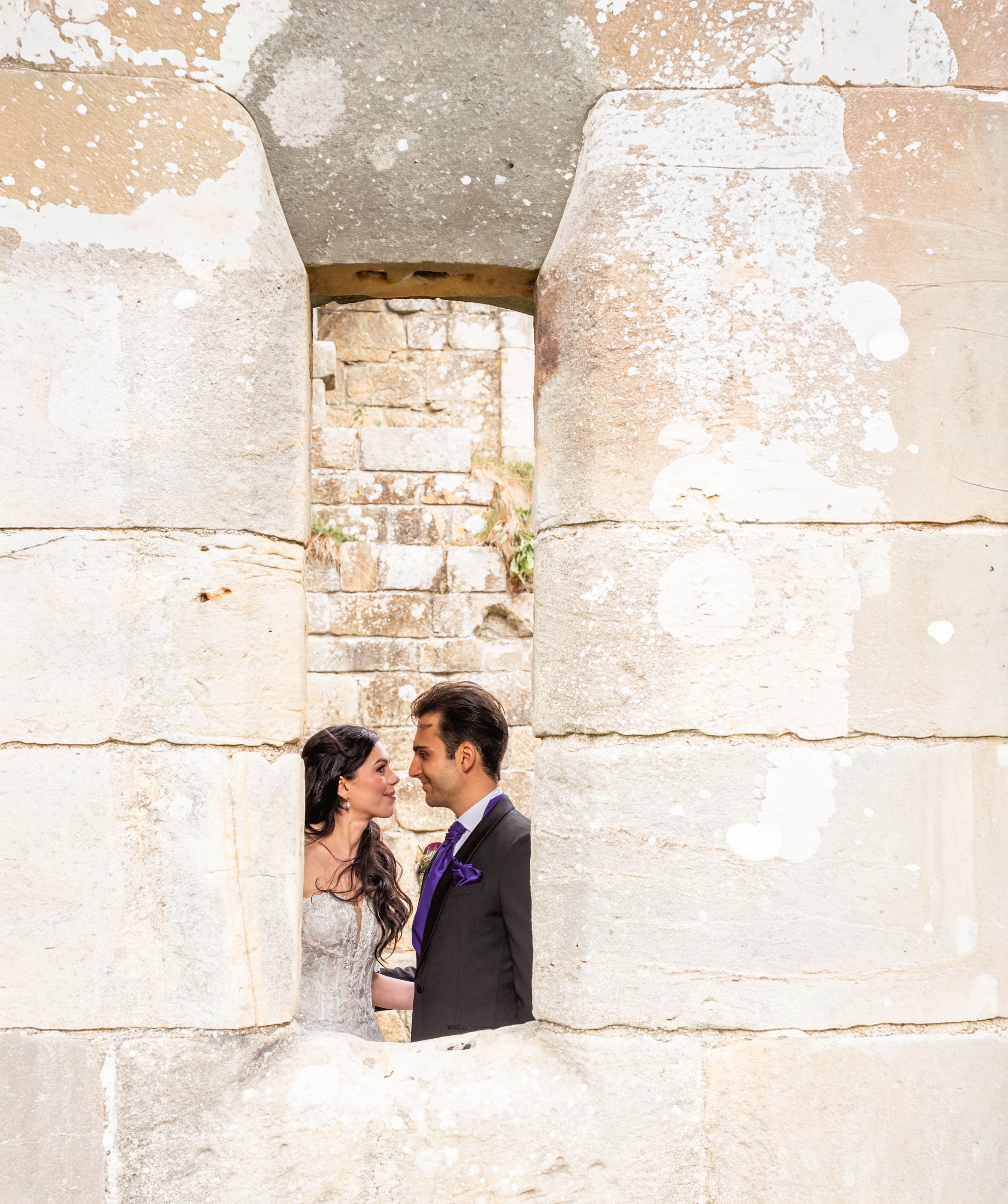 A bride and groom looking at each other through a stone opening in an ancient wall.