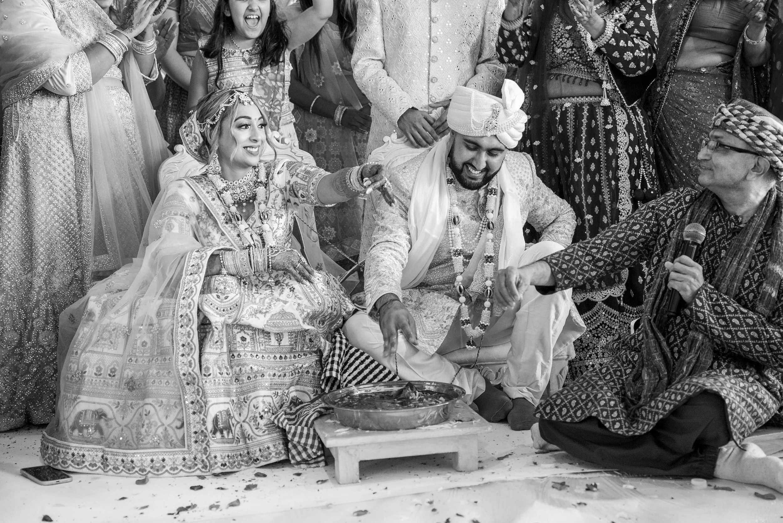 Indian wedding ceremony with bride and groom sitting on the floor. The bride is wearing traditional jewelry and attire, and the groom is wearing a turban and embroidered outfit. They are surrounded by family and friends, some smiling and celebrating.