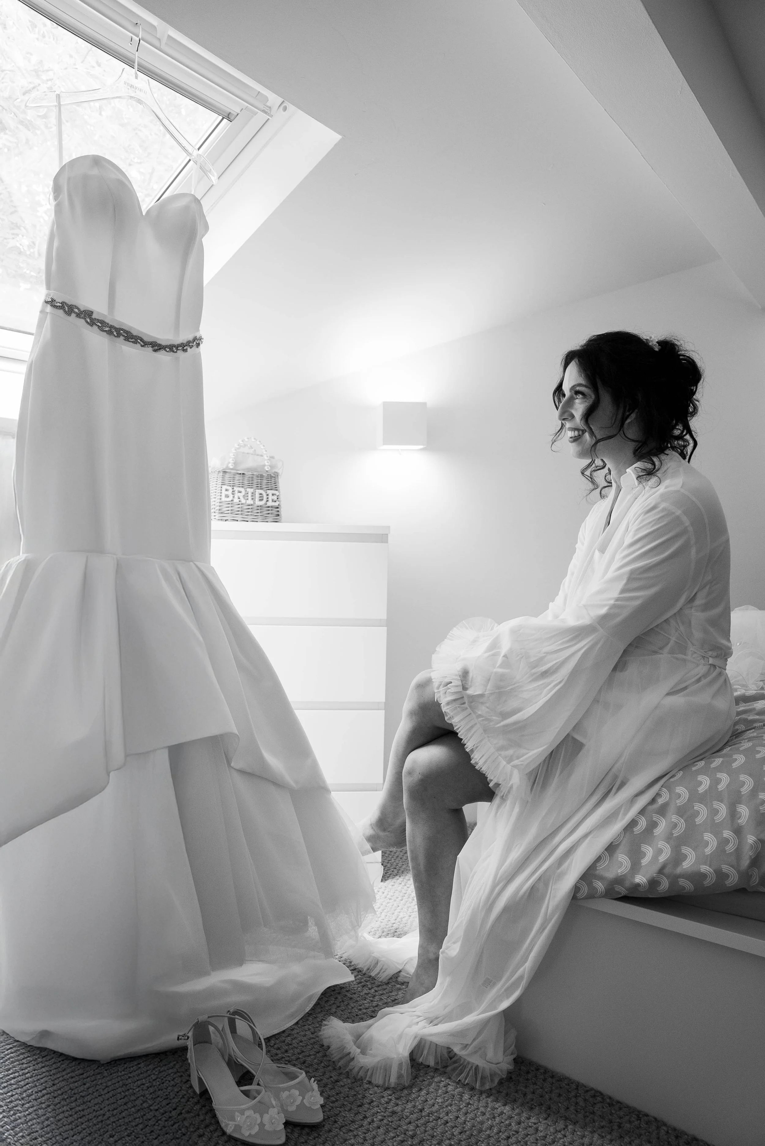 A woman sitting on her bed in a dressing gown, smiling and looking at a wedding dress hanging in front of her near a window.