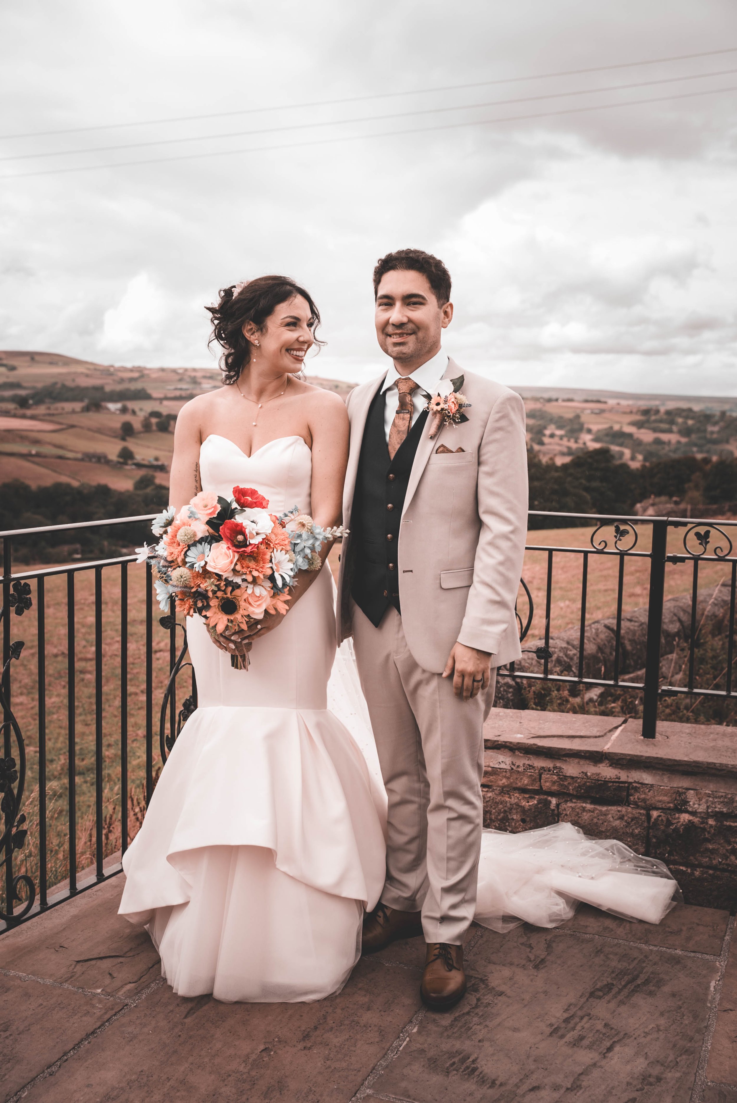 Bride and groom standing together outdoors on a cloudy day, with a scenic view of rolling hills in the background. The bride is holding a bouquet of colorful flowers and wearing a strapless white wedding gown, while the groom is dressed in a light-co