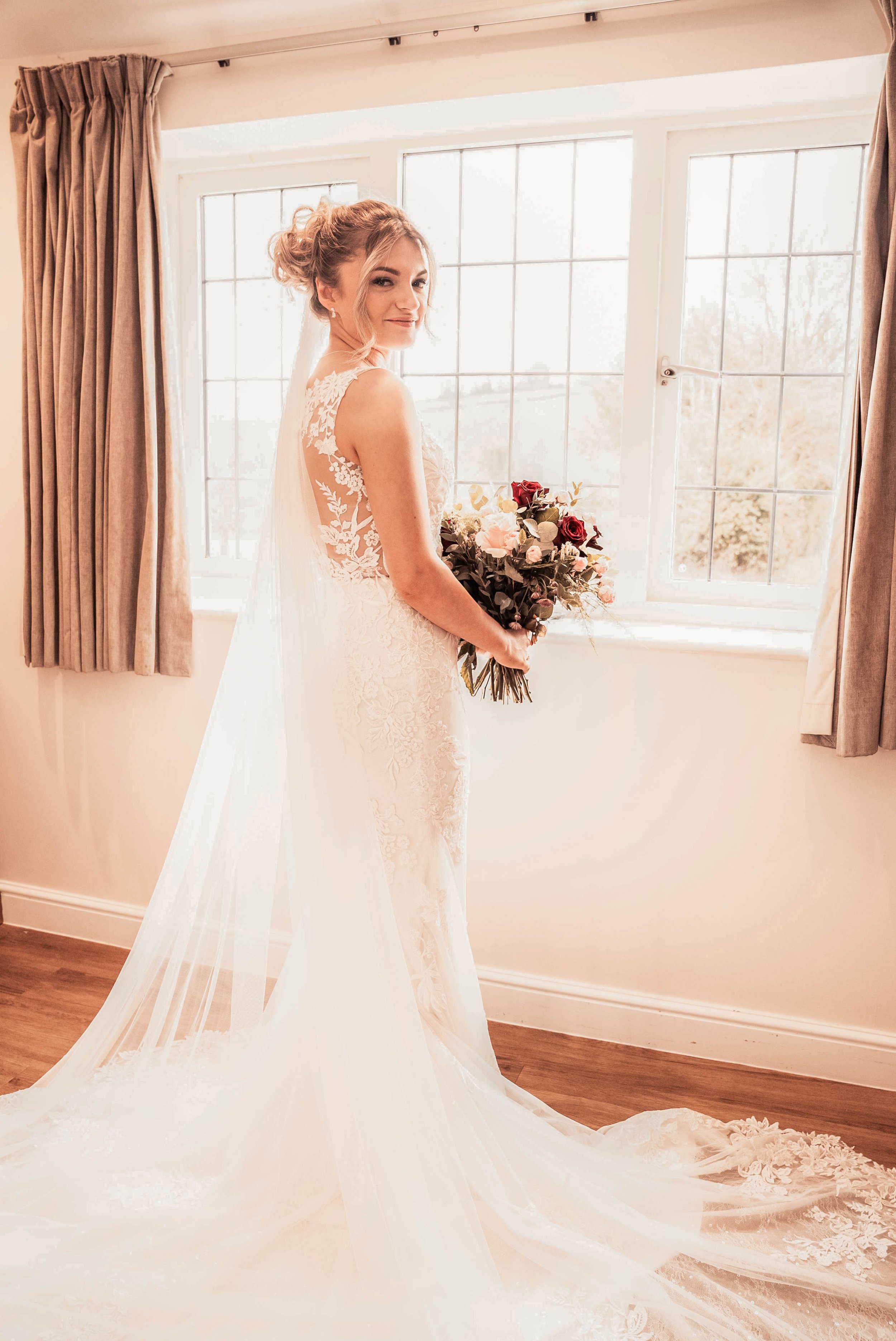 A bride in a white wedding dress holding a bouquet of pink, red, and white flowers, standing in front of a large window with beige curtains.