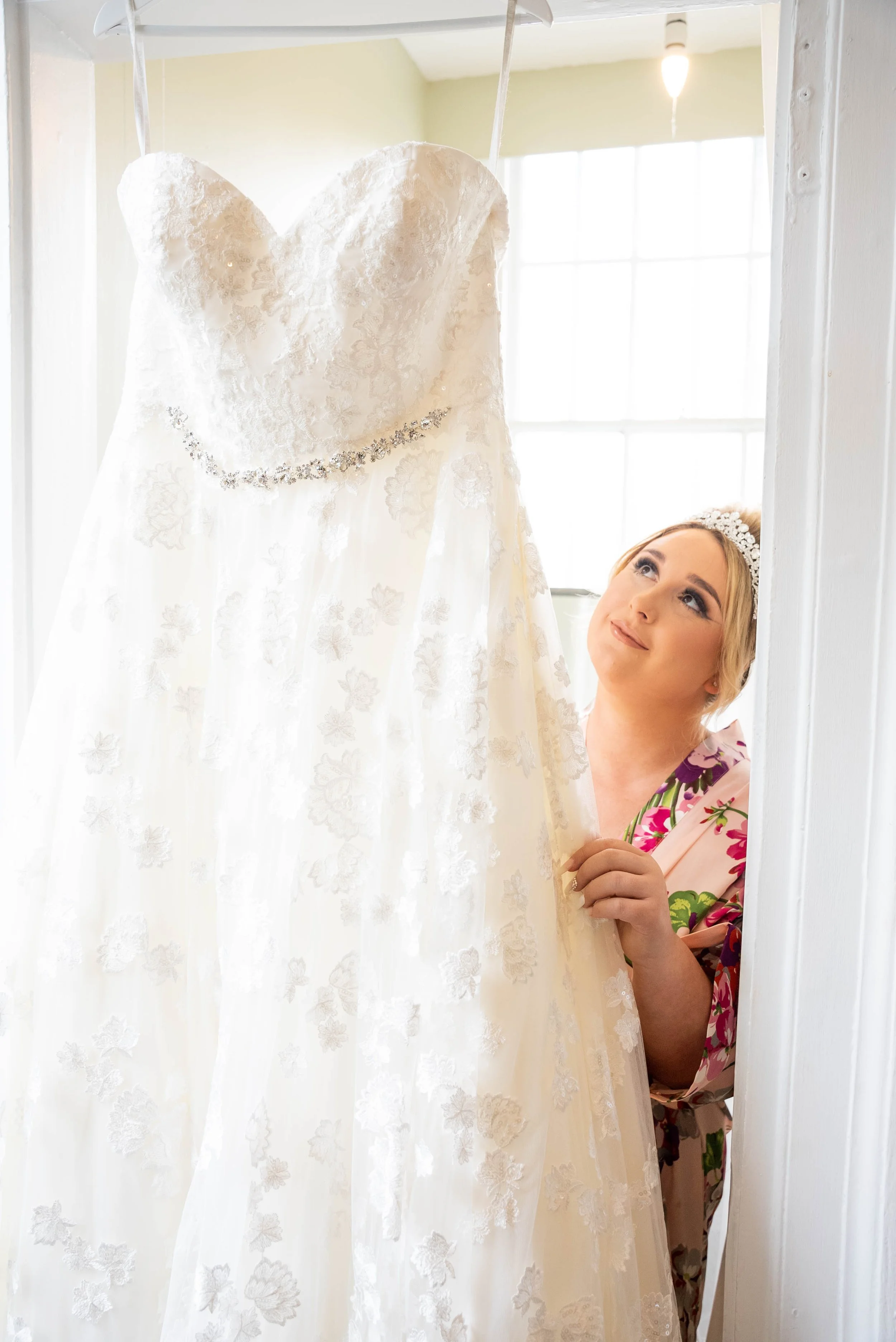 A woman with a floral headband, wearing a pink floral pajamas, looks up at a hanging white wedding dress with lace details and a crystal belt, in a bright room with a large window.