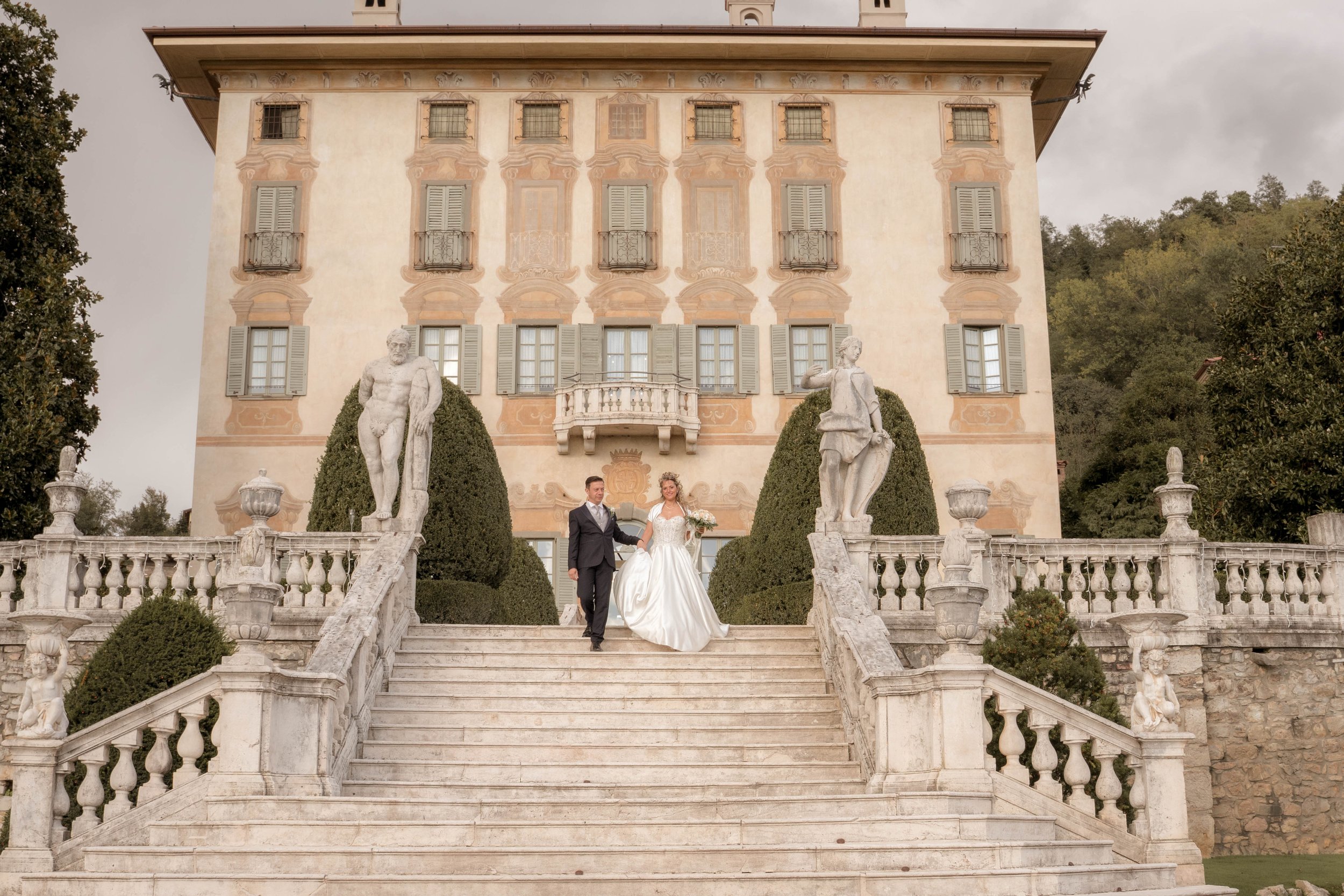 Wedding couple walking down marble stairs in front of a historic mansion with statues, greenery, and Italian-style architecture.