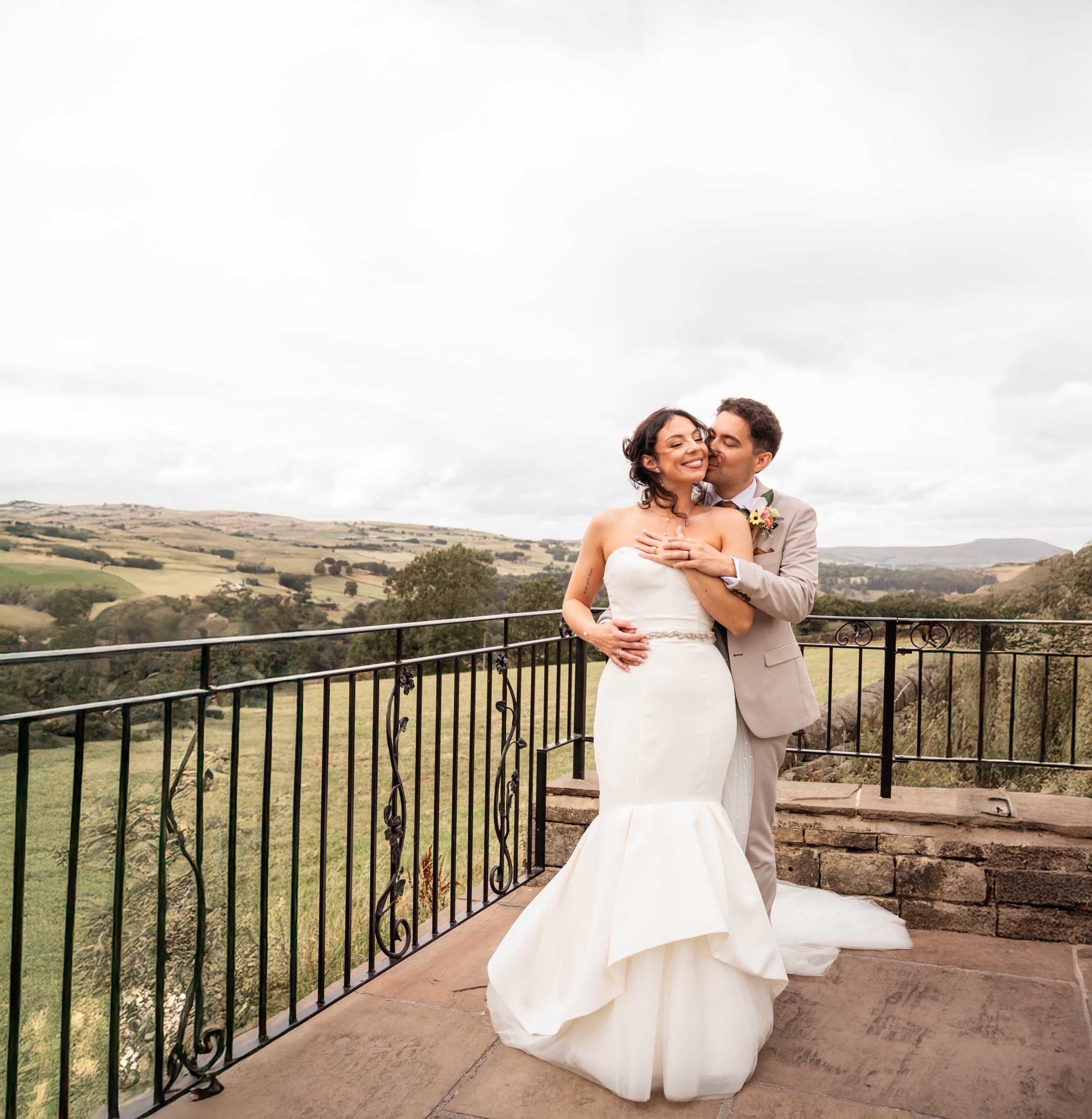 A wedding couple in wedding attire on a balcony overlooking a scenic rural landscape, with the man kissing the woman's cheek and her smiling with eyes closed.