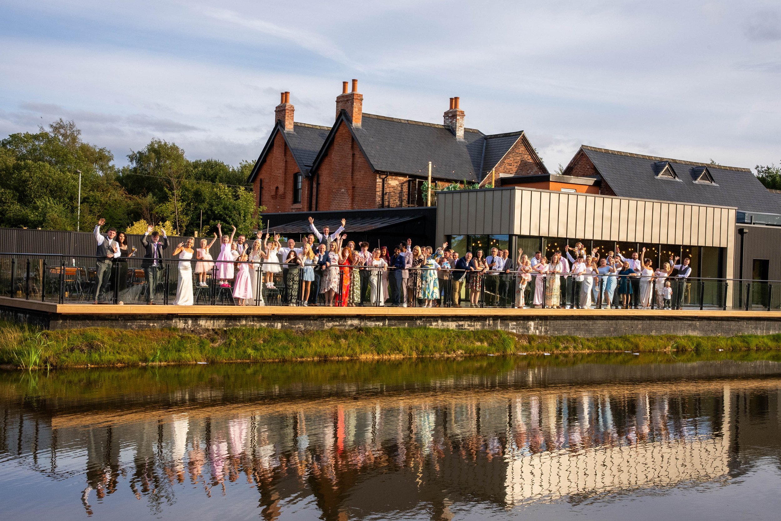 Group of people celebrating on an outdoor terrace by a river, with houses and trees in the background.