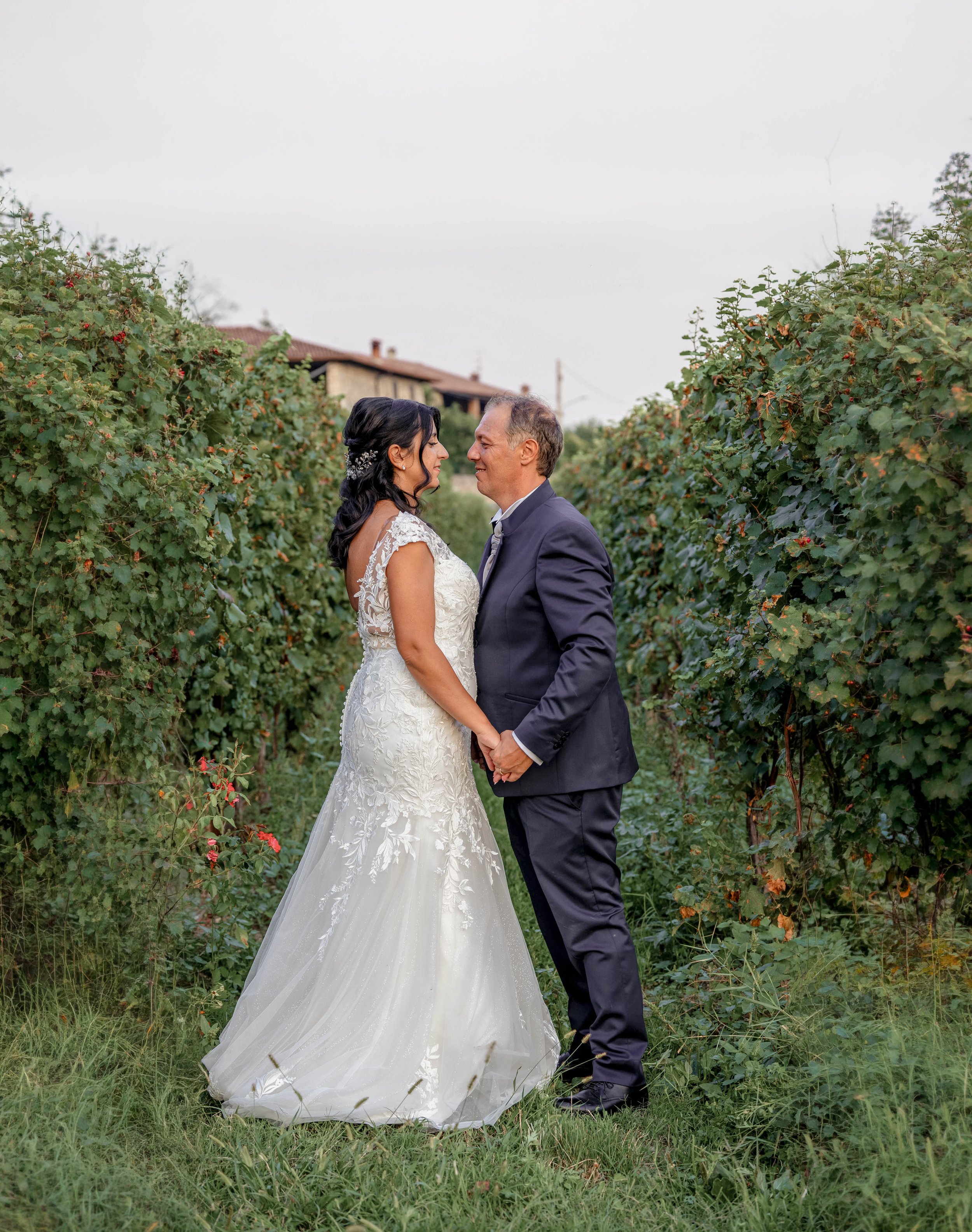 A bride and groom holding hands and smiling at each other in a vineyard with green foliage and a house in the background.