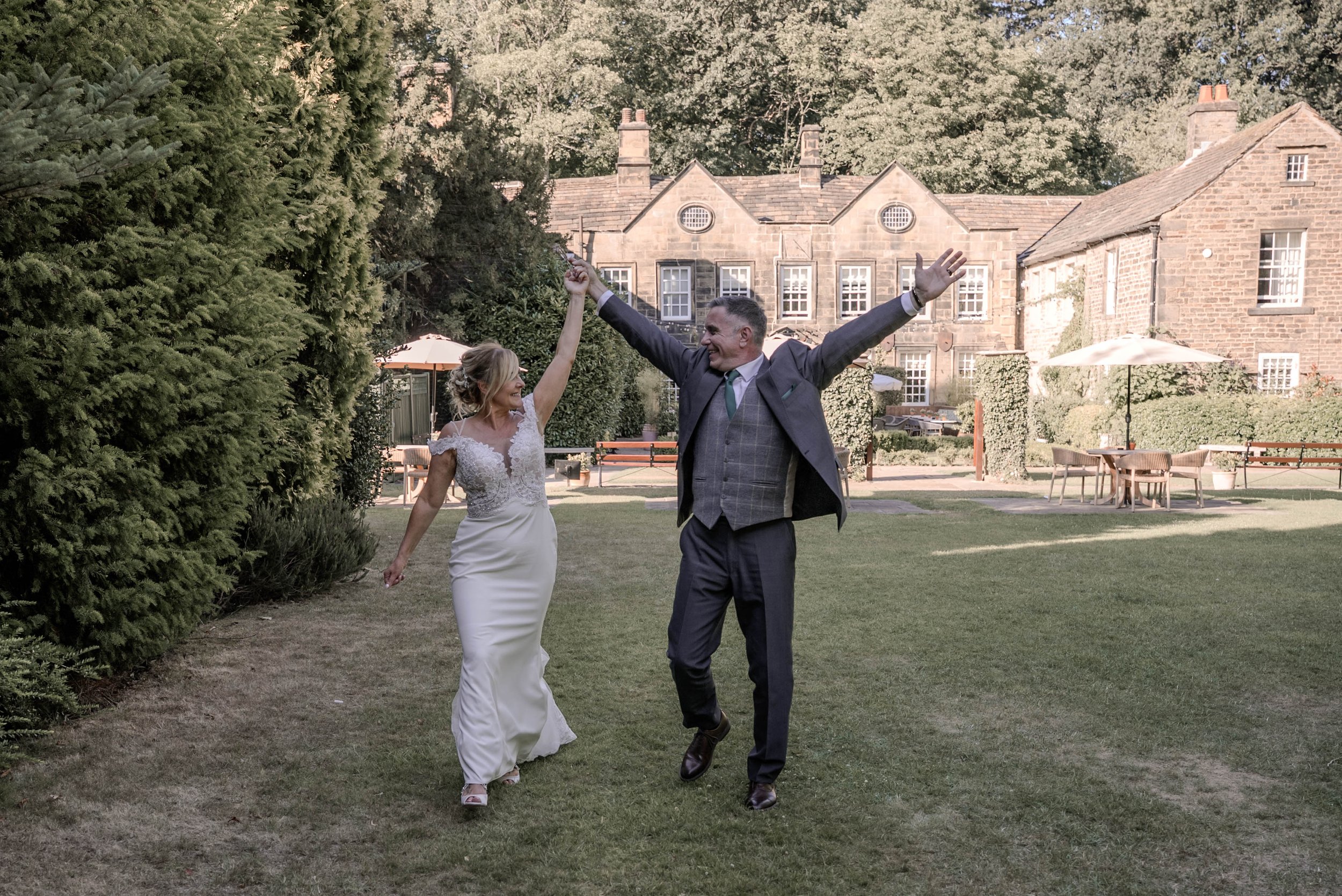 A bride and groom dancing joyfully in a garden, holding hands, with a historic stone building and outdoor tables with umbrellas in the background.