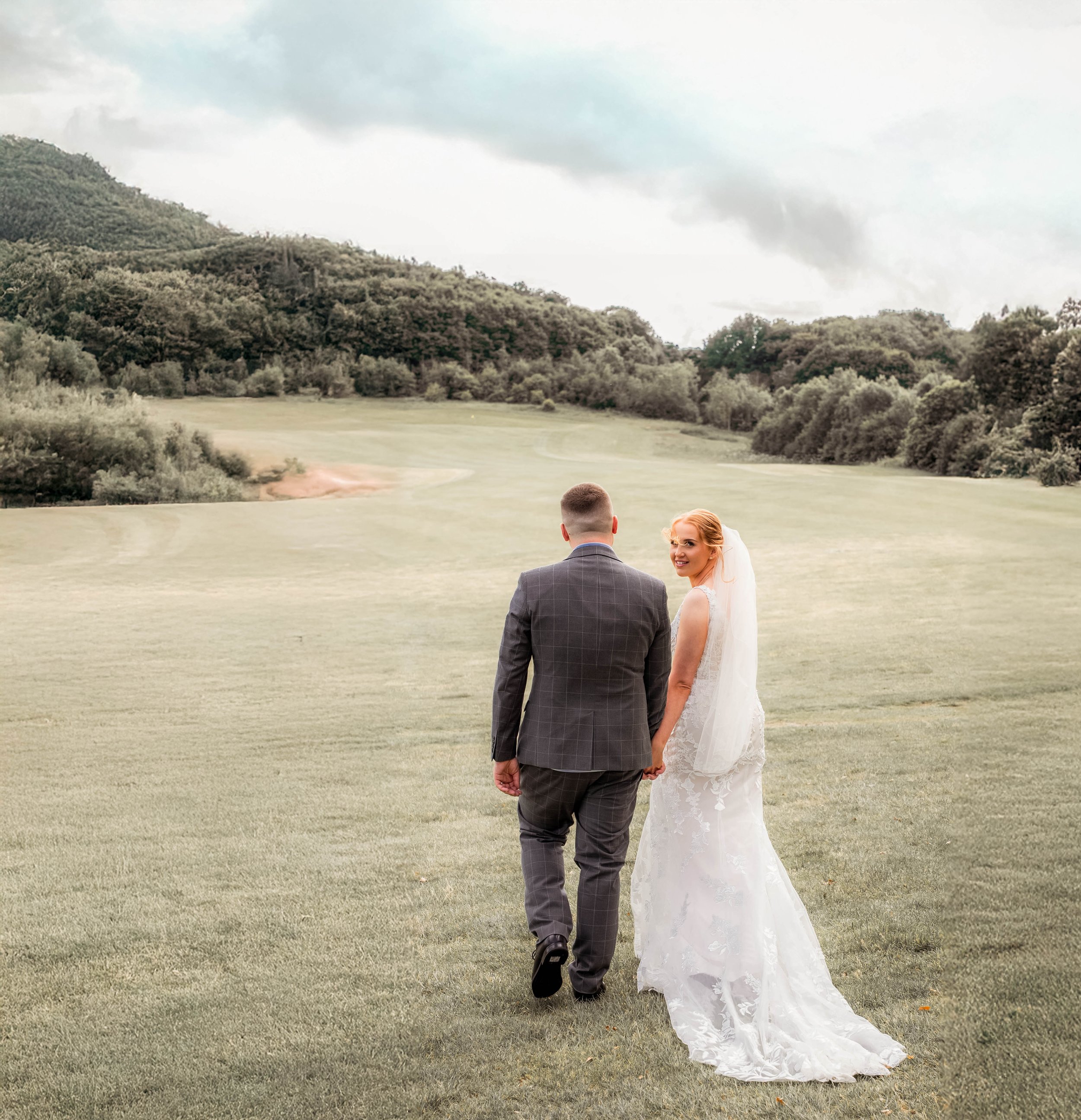 A bride and groom walking hand in hand on a golf course, holding hands, with a scenic backdrop of hills and trees under a cloudy sky.