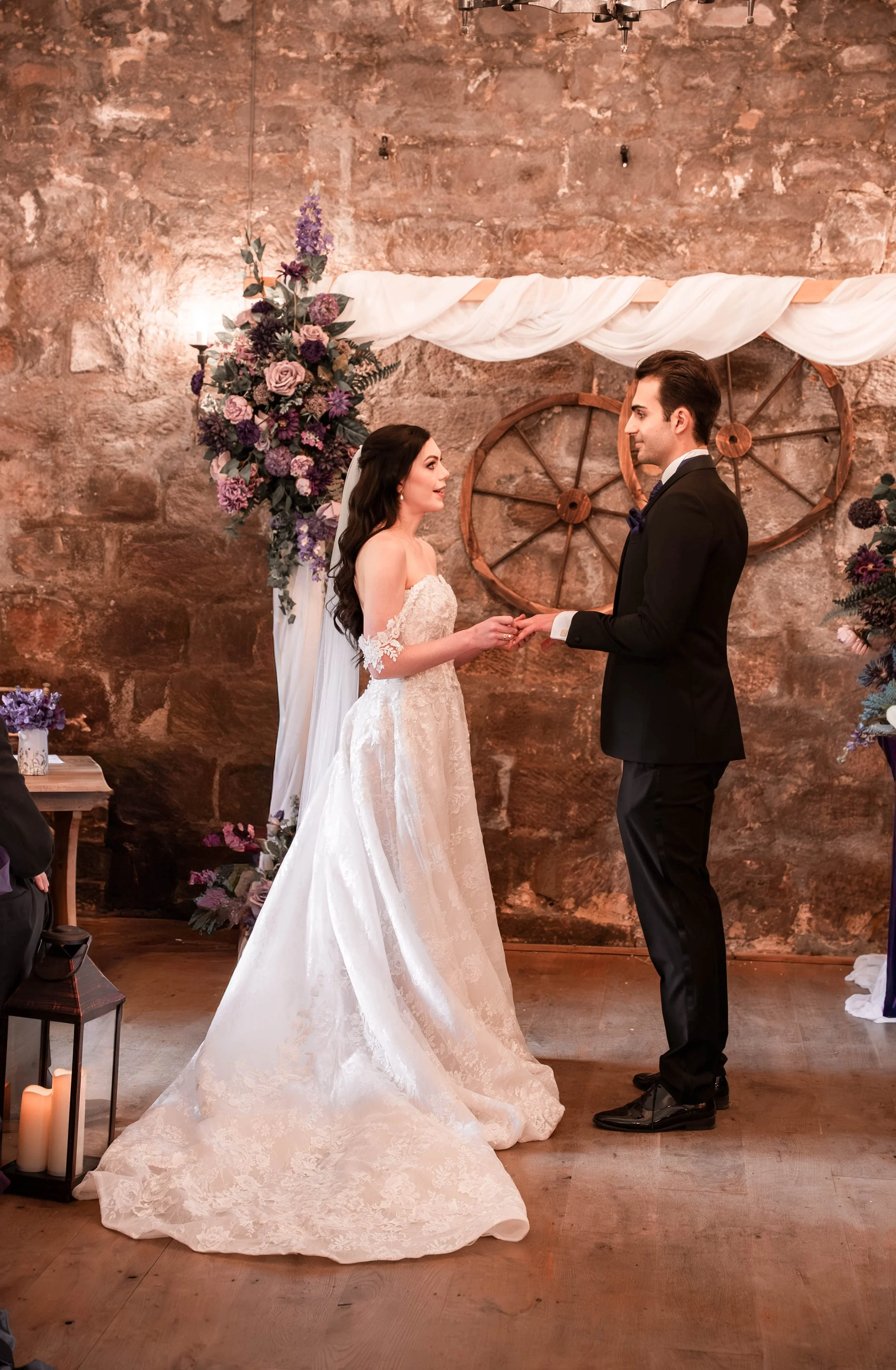 A bride and groom holding hands during their wedding ceremony inside a rustic venue with stone walls, decorative wagon wheels, and floral arrangements.