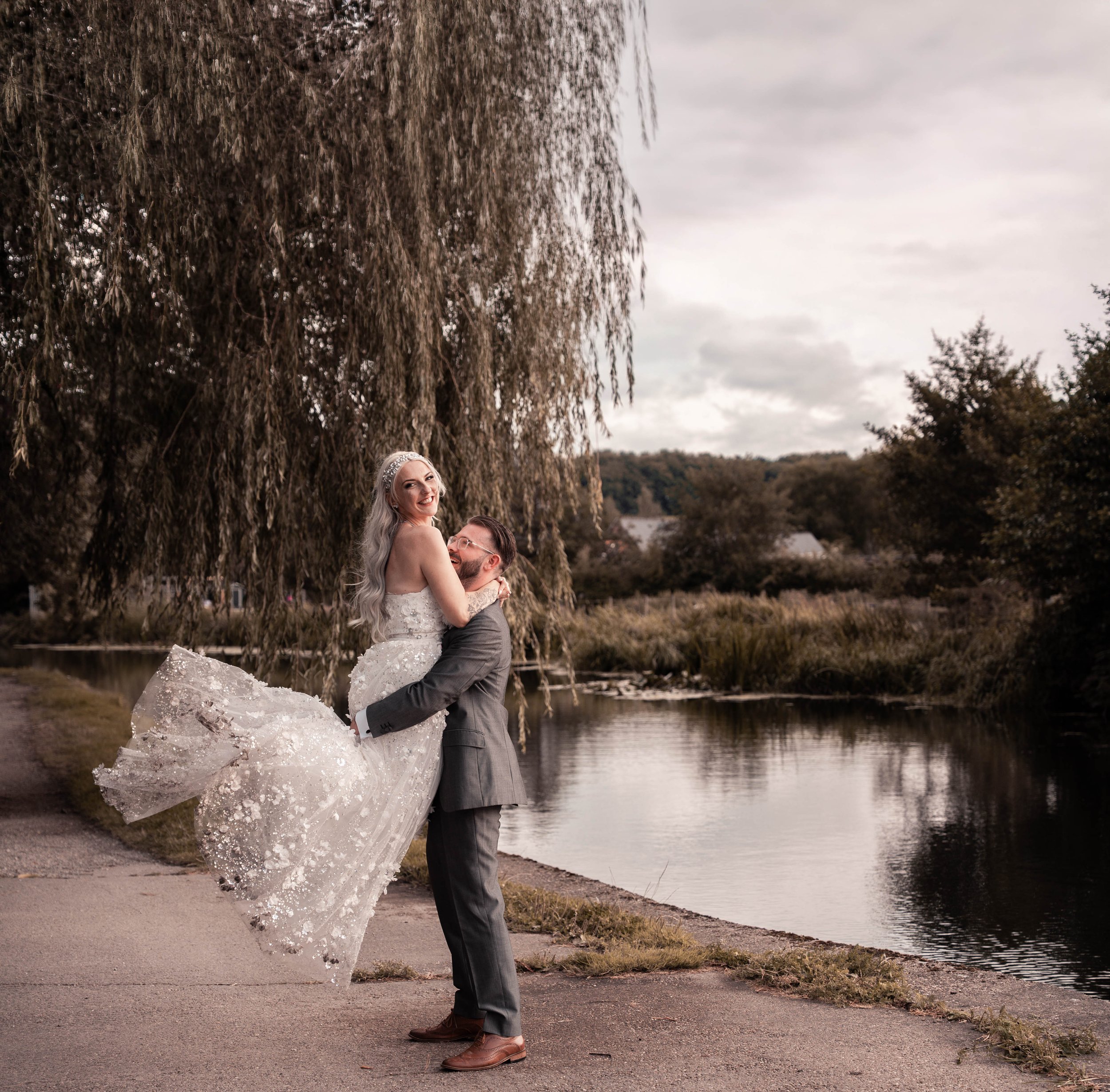 A wedding couple stands near a lake under a large weeping willow tree, with the woman wearing a white wedding dress and the man wearing a gray suit. The woman is smiling at the camera while the man embraces her. The scene is outdoors with cloudy skie