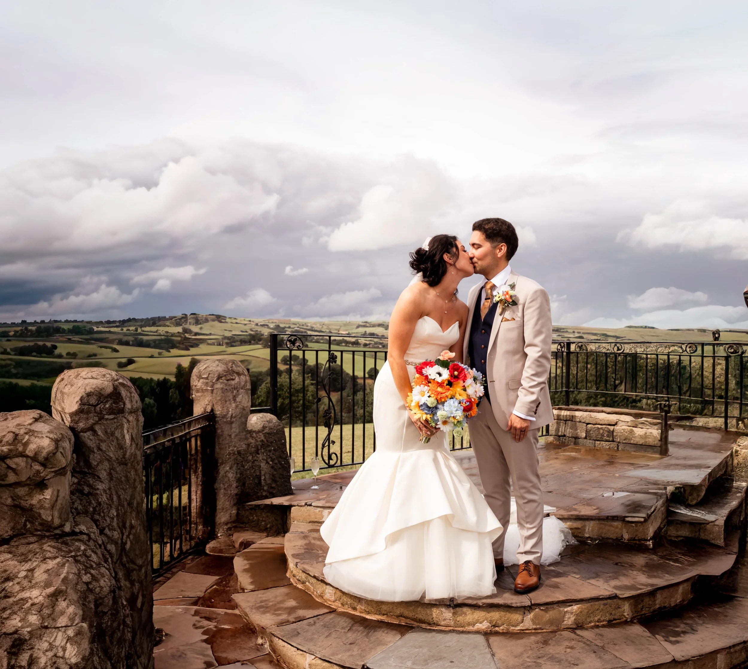 A newlywed couple kisses on a stone platform with a scenic landscape of rolling hills and a cloudy sky in the background. The bride is holding a colorful bouquet, and the groom is dressed in a light-colored suit.
