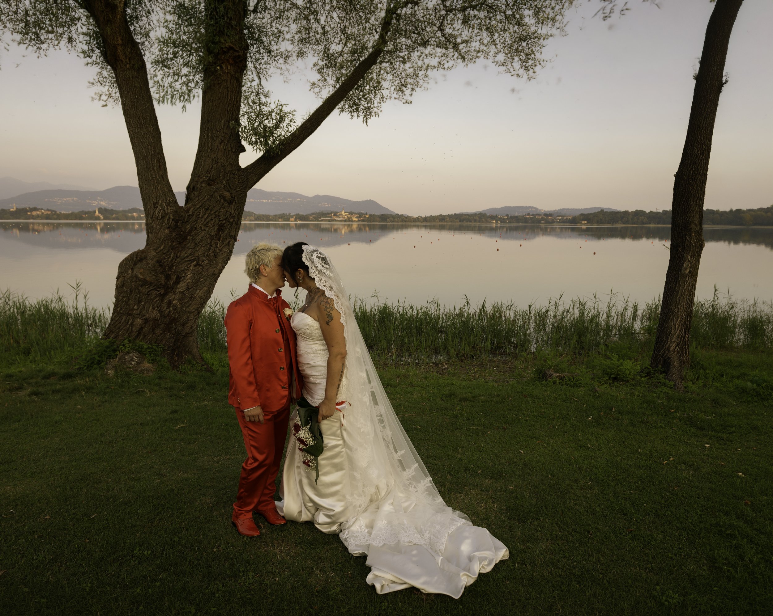 A wedding couple stands close together, under a tree by a lake at sunset. The groom wears a red suit and the bride wears a white wedding gown with a lace veil, holding a bouquet, with their foreheads touching in an intimate pose.