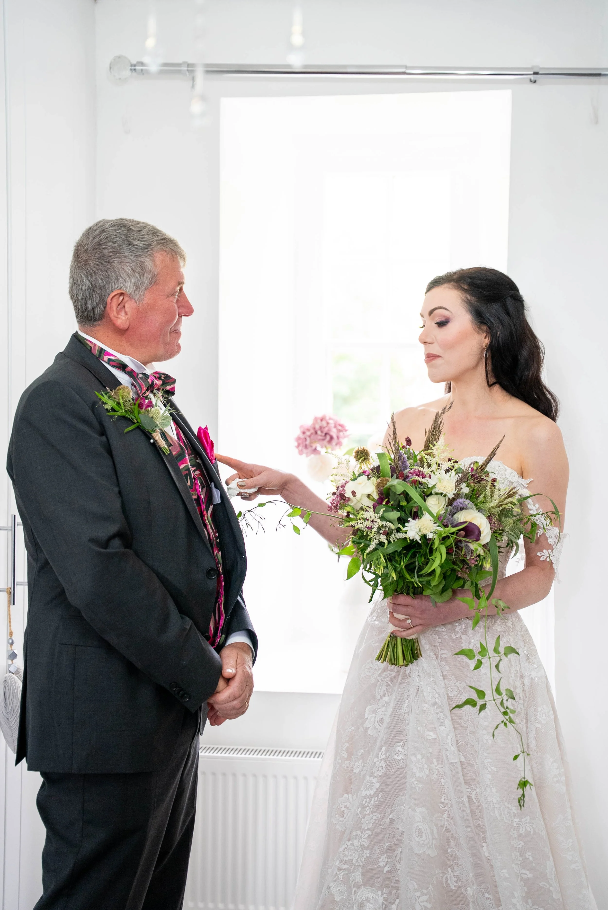 A bride in a lace wedding gown holding a bouquet of flowers stands facing an older man in a suit. The man wears a boutonniere and a colorful striped tie. They are in a well-lit room, possibly during a moment of wedding vows or a special exchange.