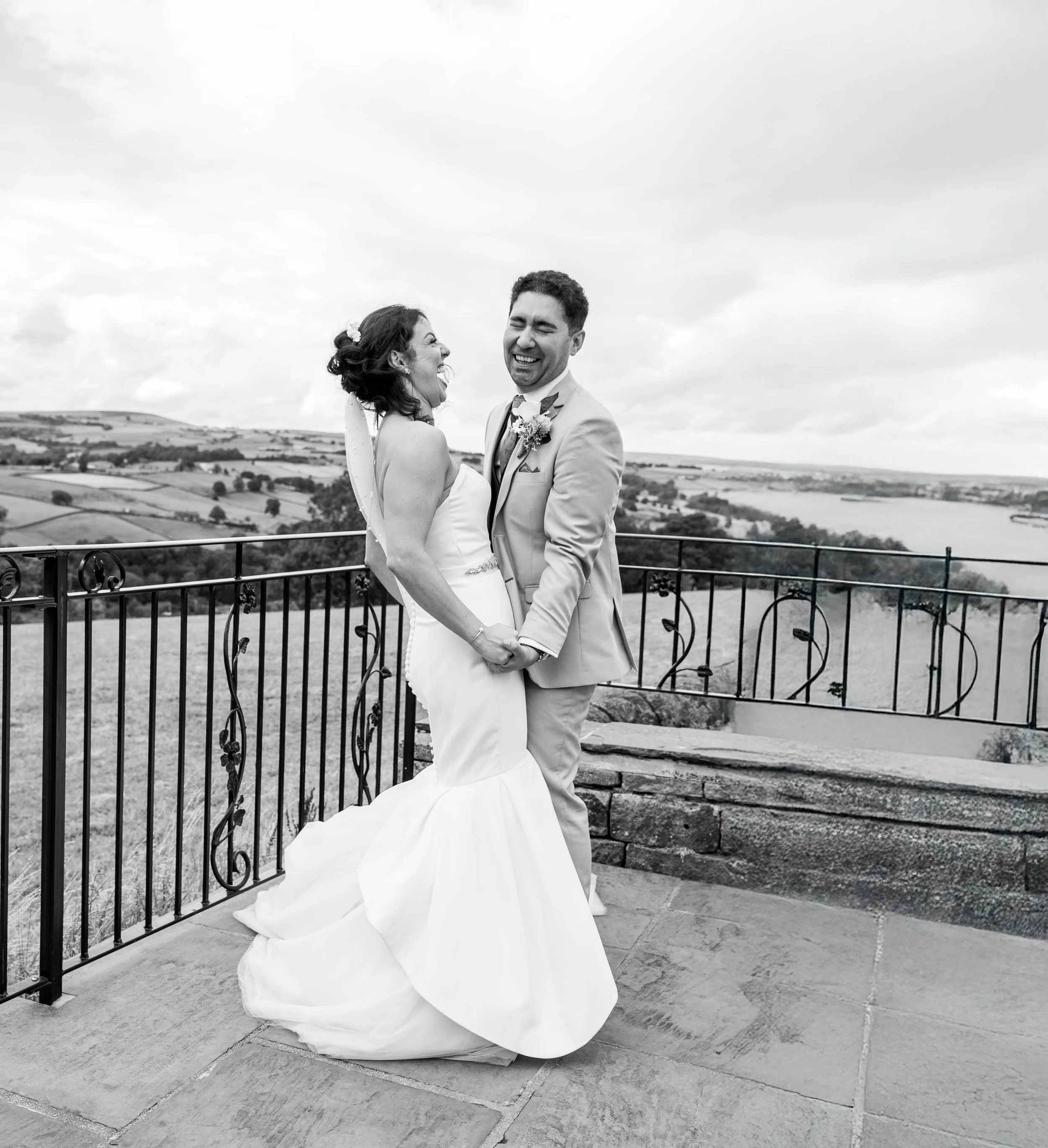 Black and white photo of a smiling bride and groom holding hands on a balcony with a scenic countryside in the background during their wedding.