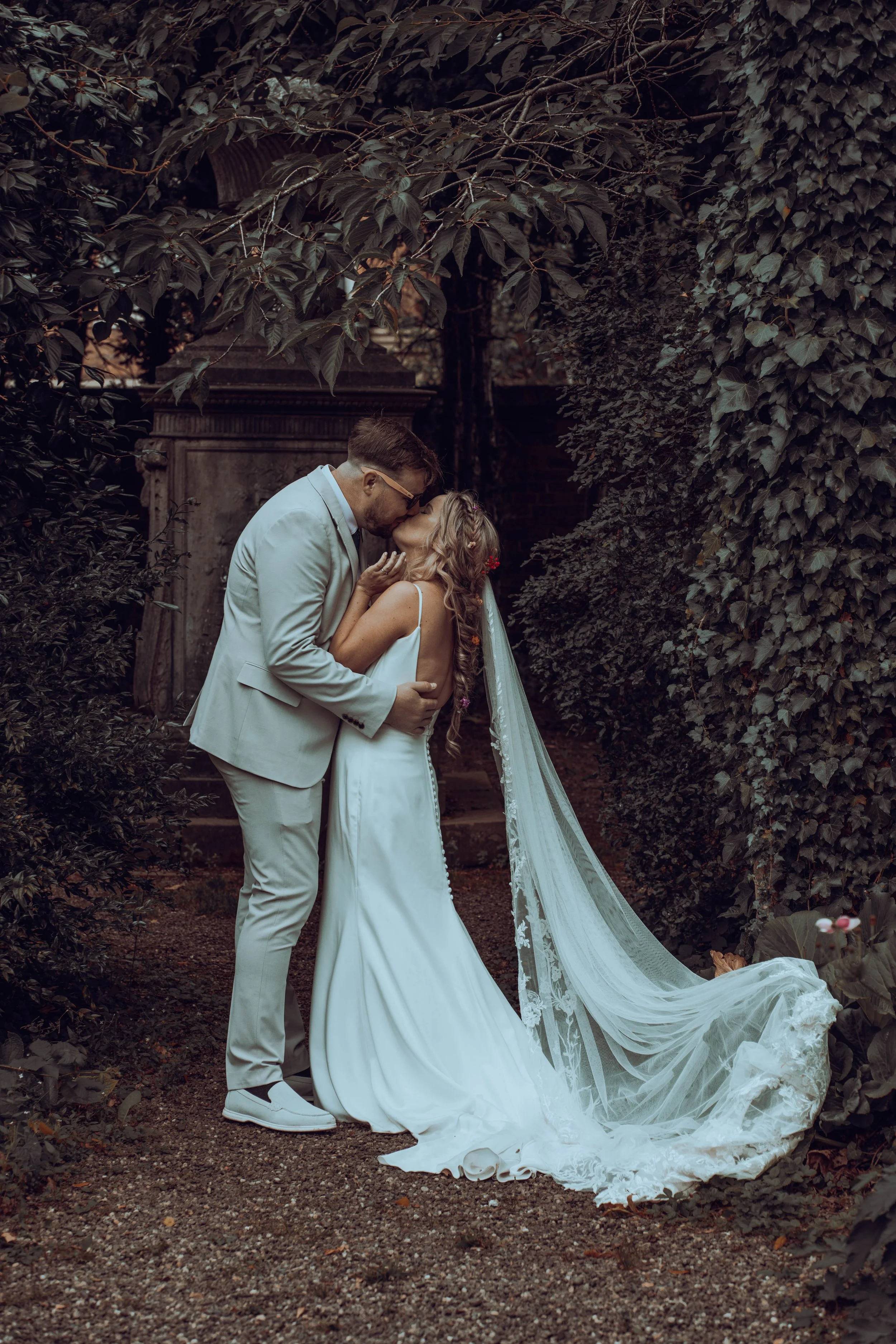 A couple in wedding attire sharing a kiss outdoors surrounded by lush foliage.