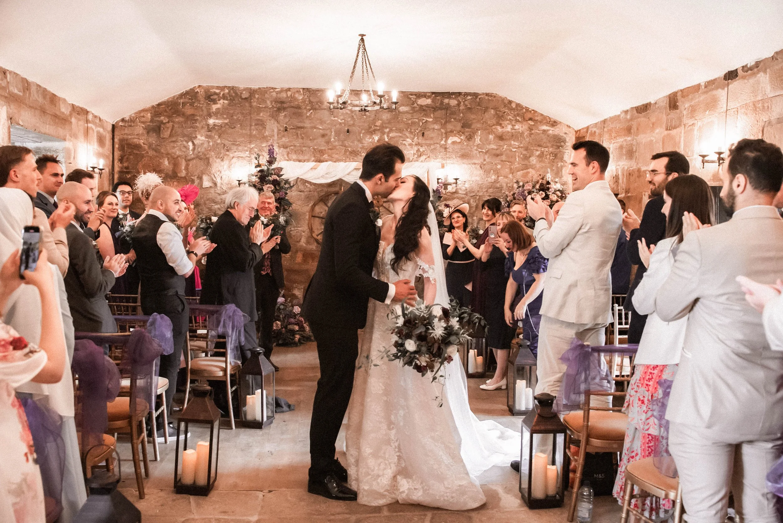 A wedding ceremony with a bride and groom kissing, surrounded by a seated and standing guests, in a rustic stone-walled venue decorated with floral arrangements, candles, and lanterns.