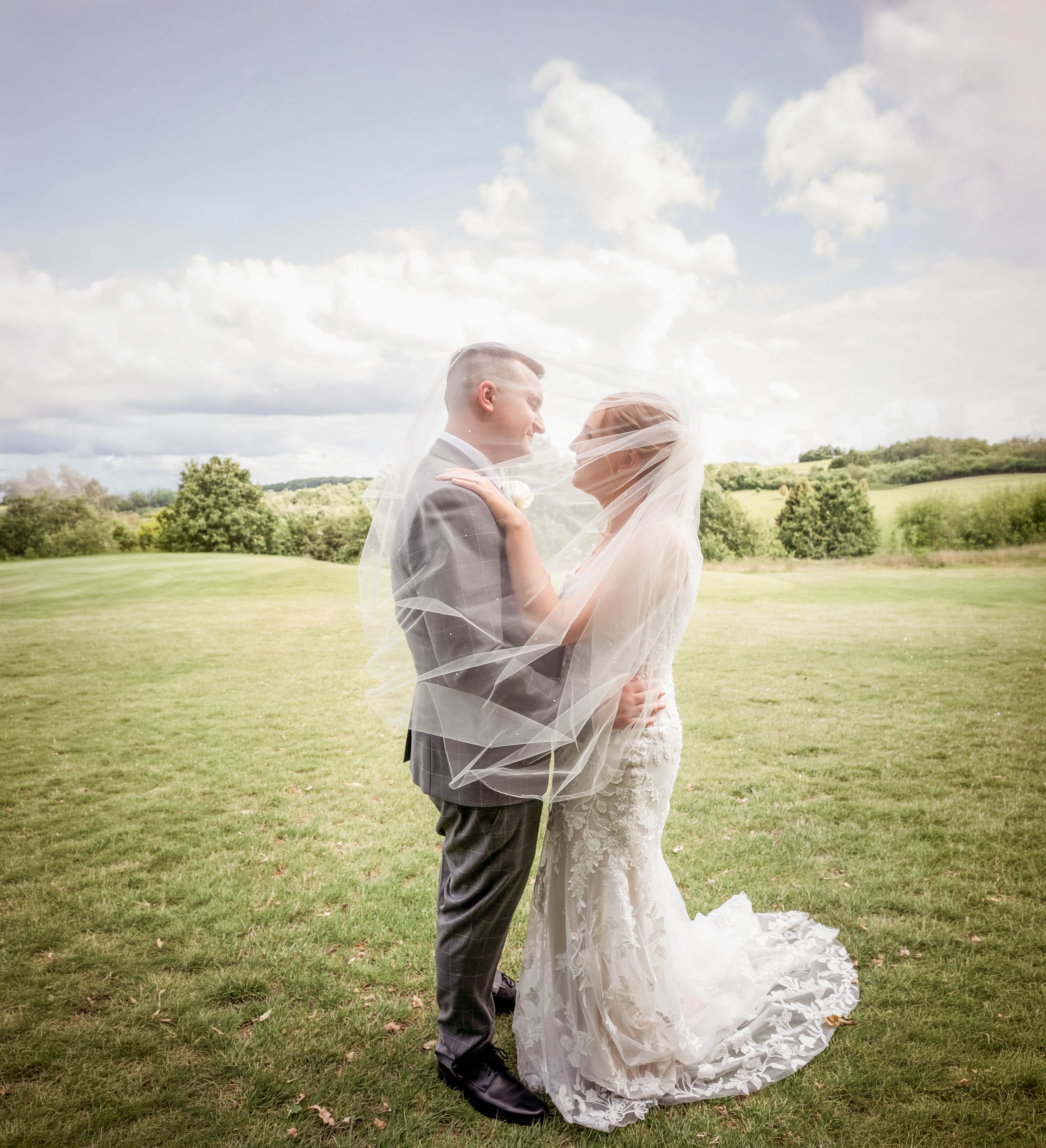 A bride and groom are standing close together outdoors on a grassy field, smiling at each other, with a veil draped over both of them, during their wedding photo shoot.