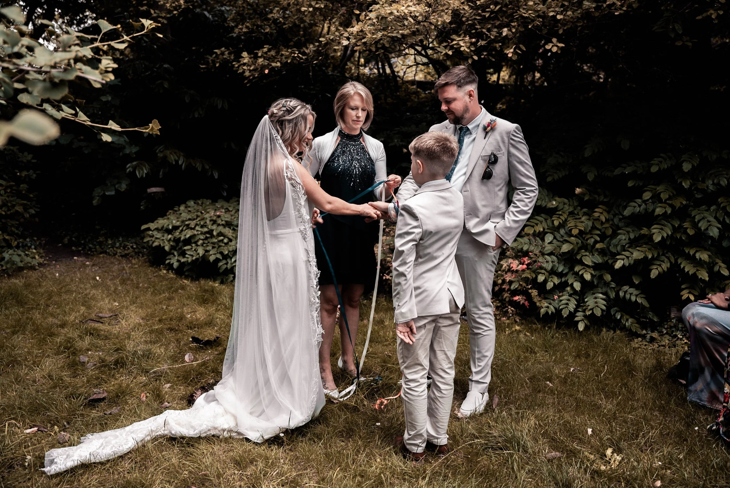A bride and groom participating in a wedding ceremony outdoors, exchanging rings, with an officiant and a young boy present, in a lush garden setting.