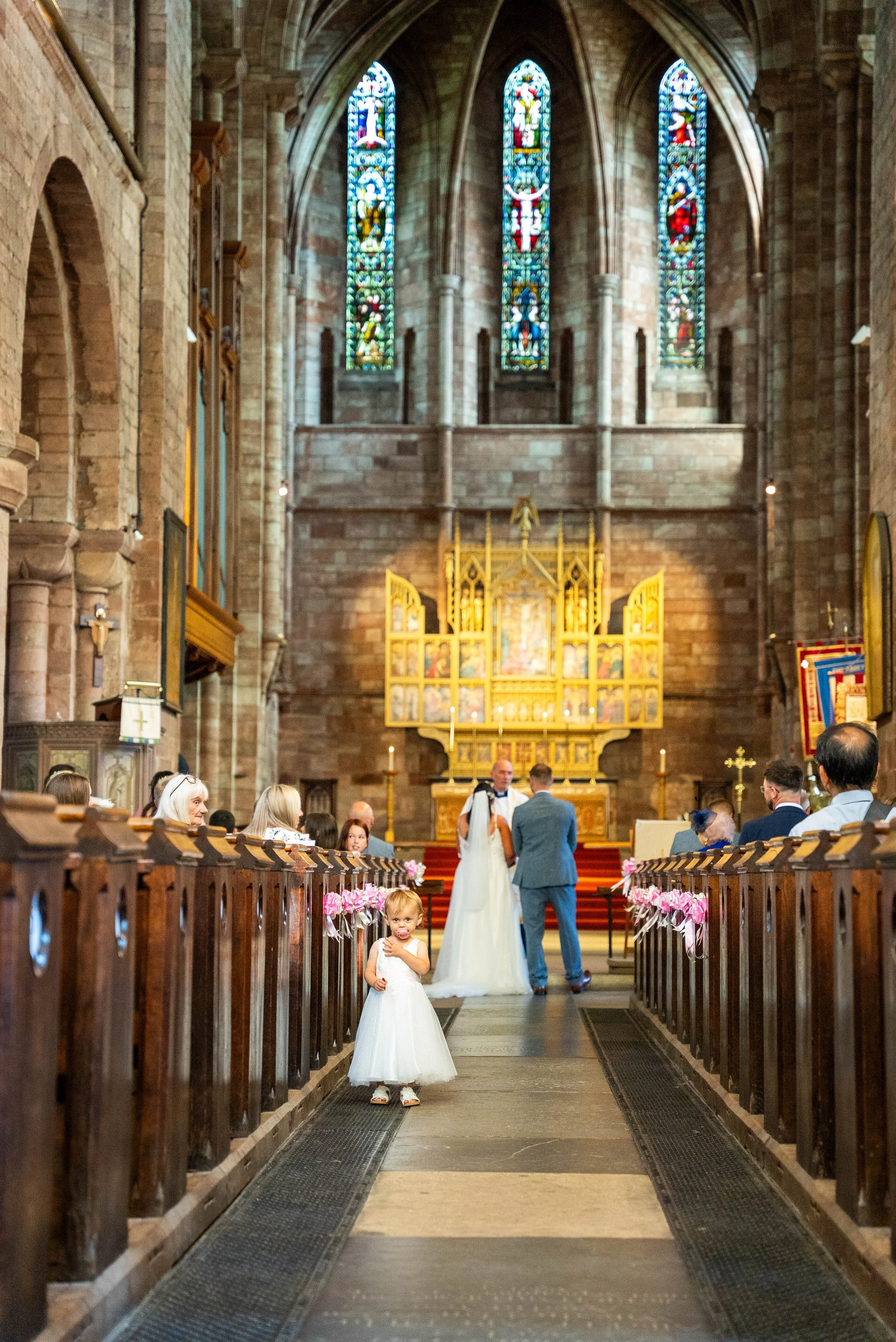 A wedding ceremony inside a church with stained glass windows and an ornate altar. A girl in a white dress stands in the aisle looking at the camera. Guests are seated on wooden pews, and two people are standing at the altar.