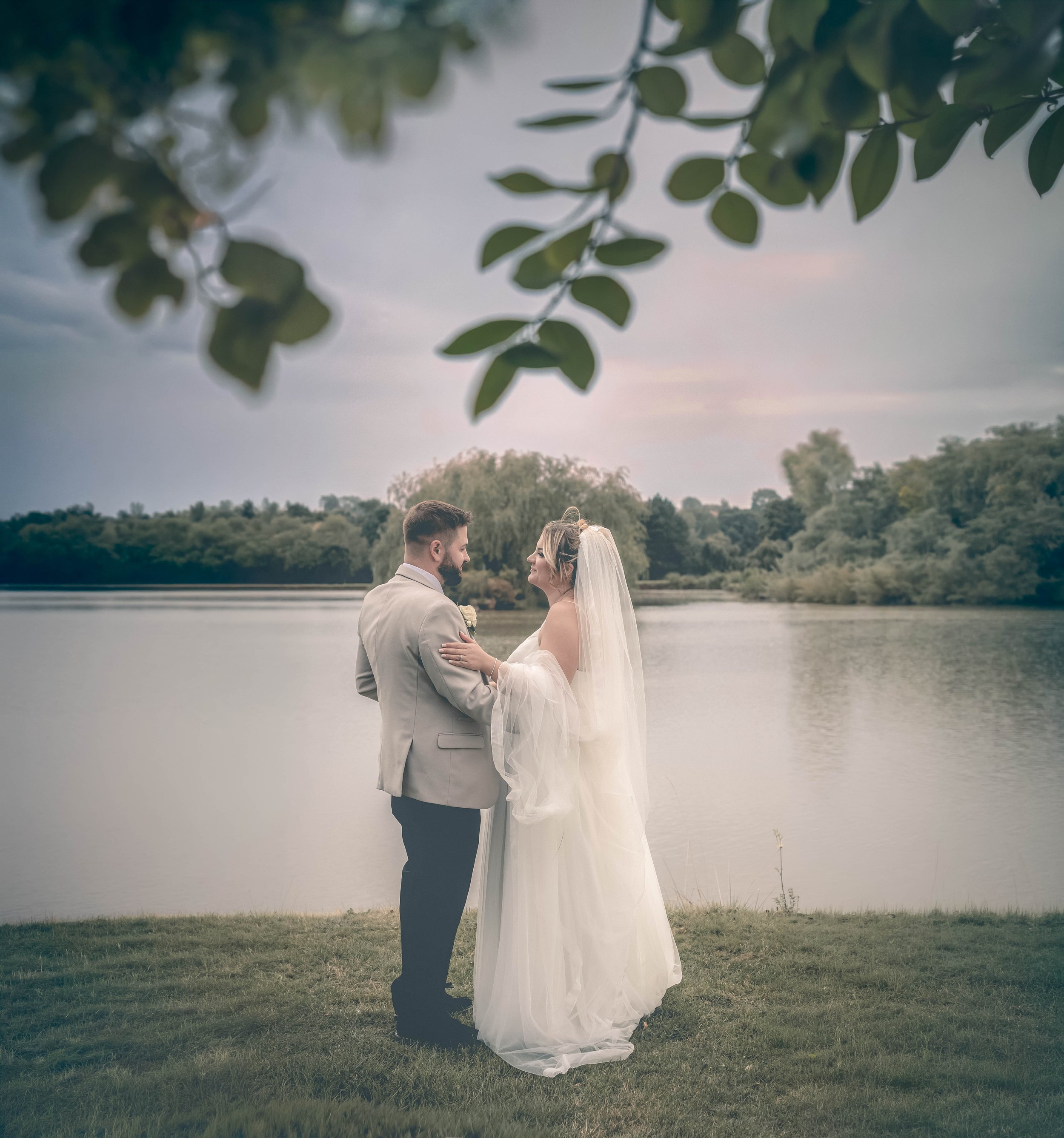 A bride and groom holding hands by a lake during sunset, with a leafy tree branch in the foreground.