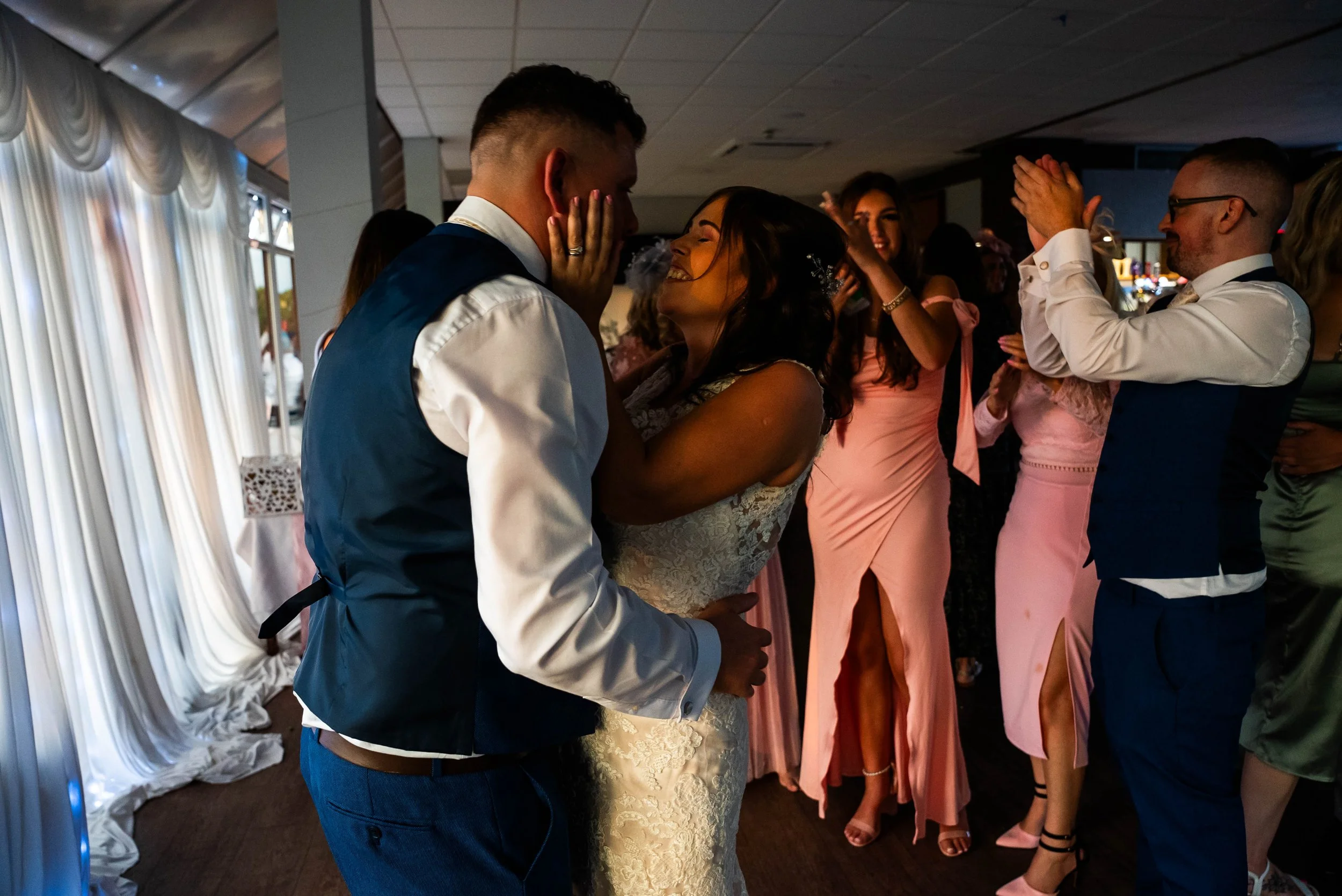 A bride and groom sharing a joyful moment on their wedding dance floor, surrounded by friends in elegant dresses and suits, with curtains and soft lighting in the background.