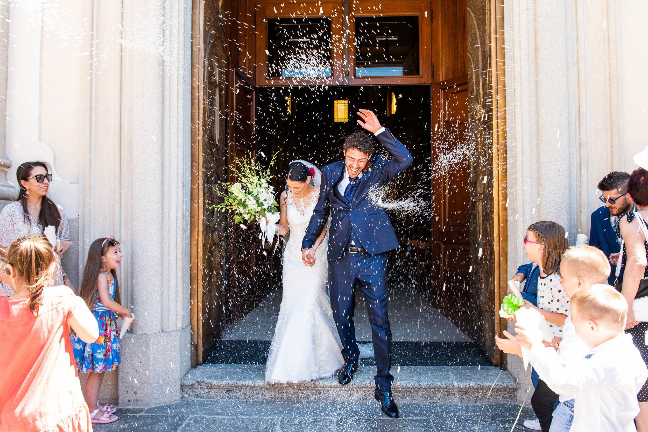 A bride and groom celebrating outside a church after their wedding, surrounded by children and adults, with confetti falling as they walk out of the church.