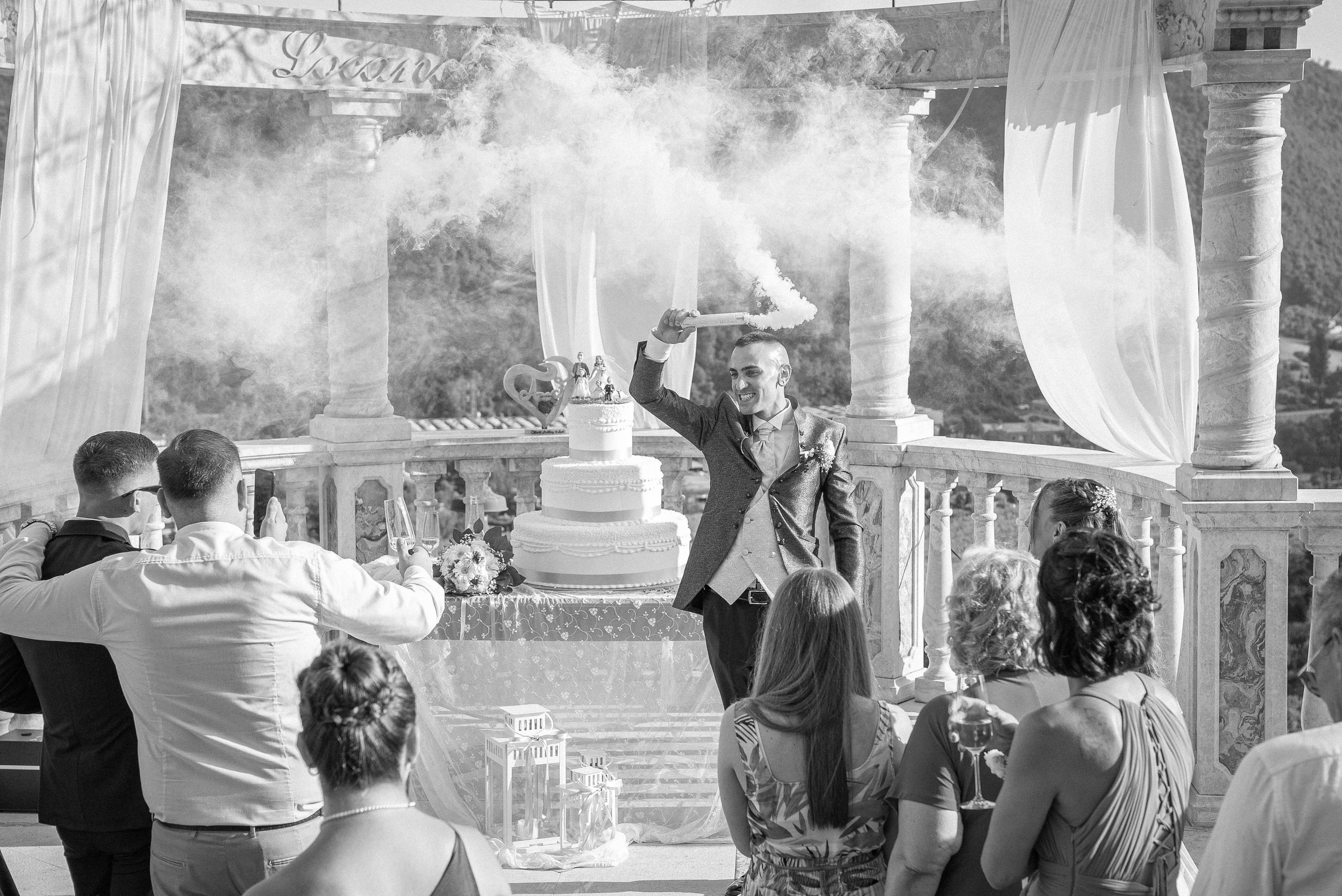 A wedding celebration with guests watching the bride and groom, with the groom holding a smoke grenade, at an outdoor venue with columns and drapery.