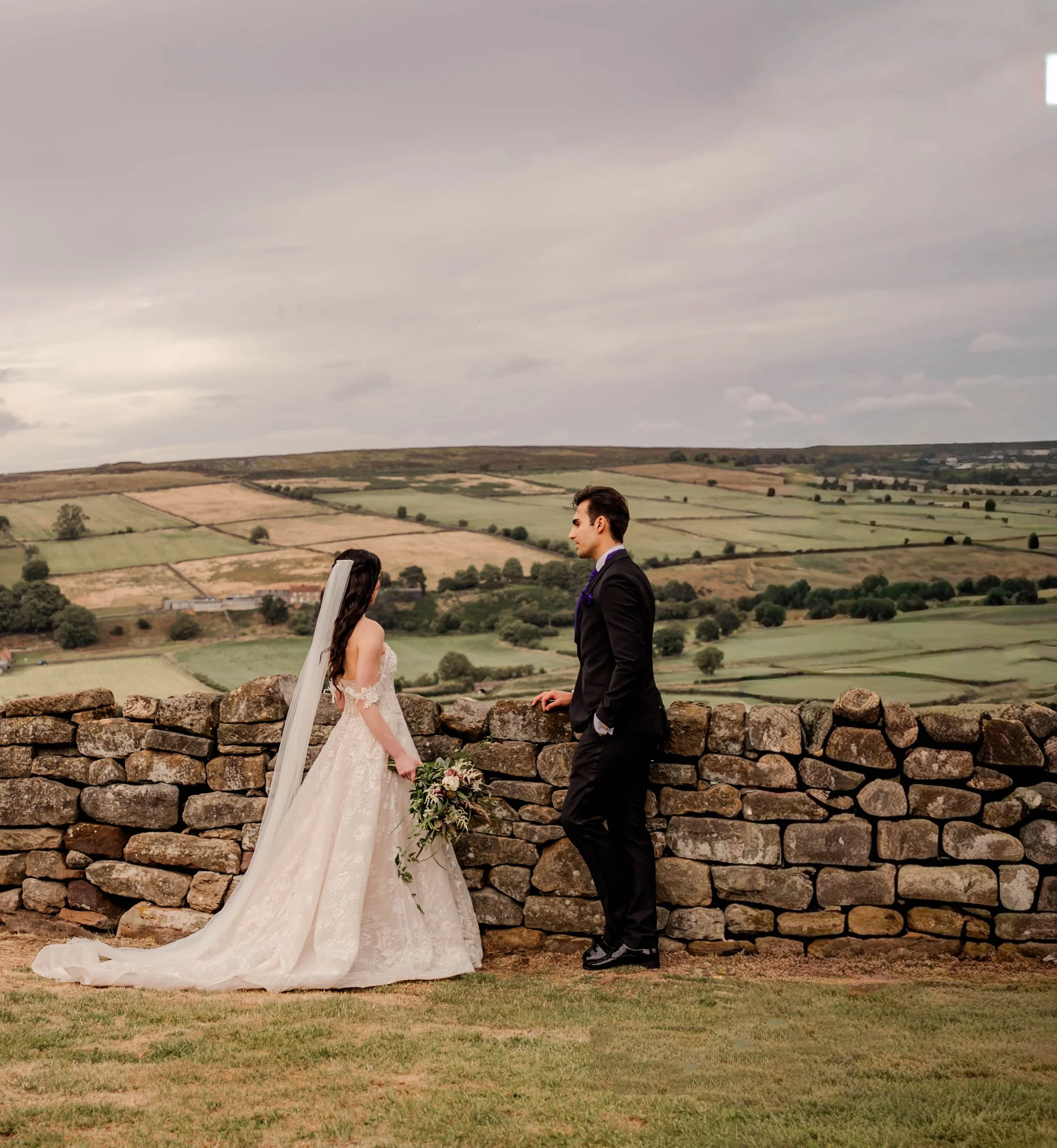 Bride and groom standing outdoors by a stone wall with a countryside landscape in the background, during their wedding.