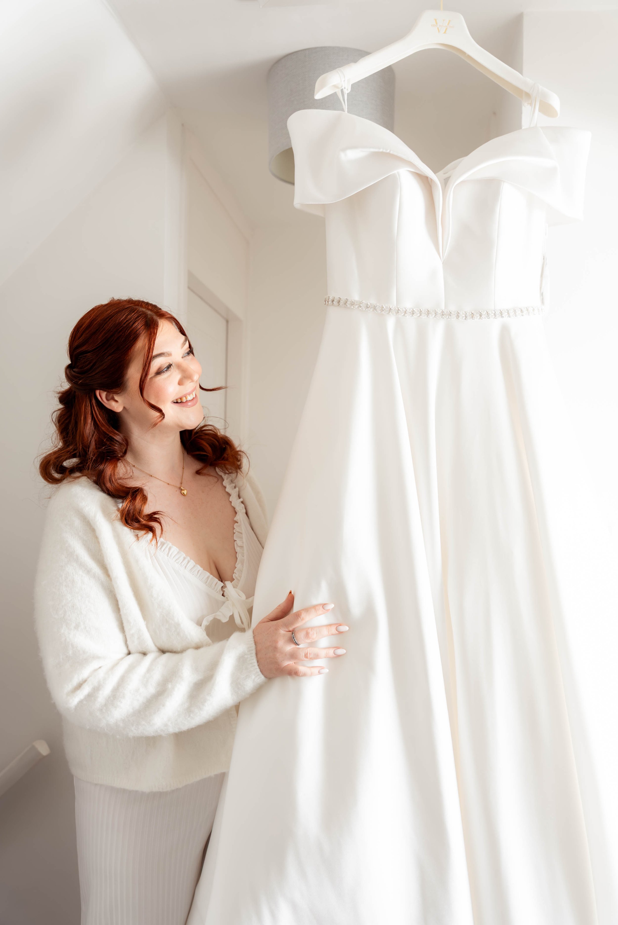 A woman with red hair smiling and looking at a wedding dress hanging in front of her.