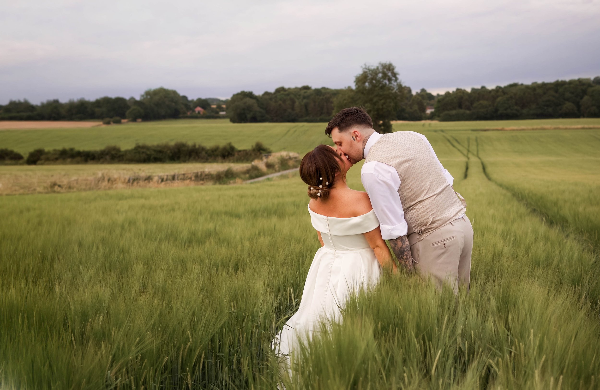 A couple dressed in wedding attire sharing a kiss in a green field with rolling hills and trees in the background under a cloudy sky.