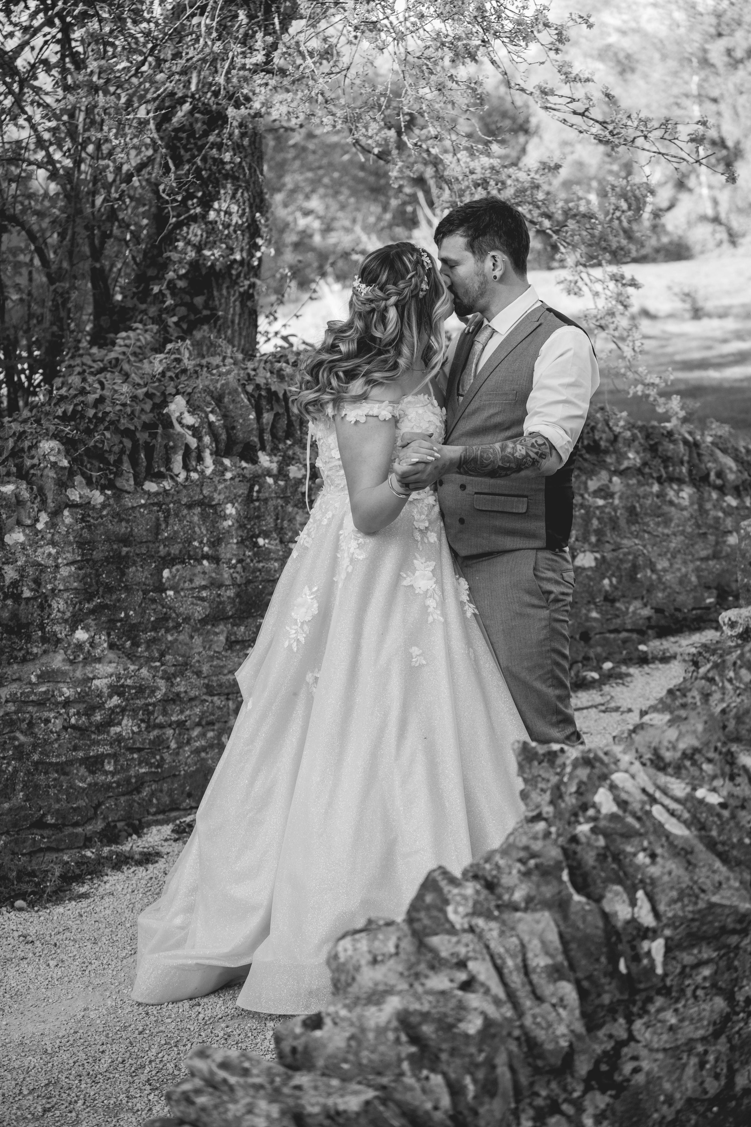 A black and white photograph of a couple kissing outdoors. The woman wears a wedding gown with floral details and a braid with flowers in her hair. The man wears a vest over a shirt with rolled-up sleeves, revealing tattooed arms. They stand close to