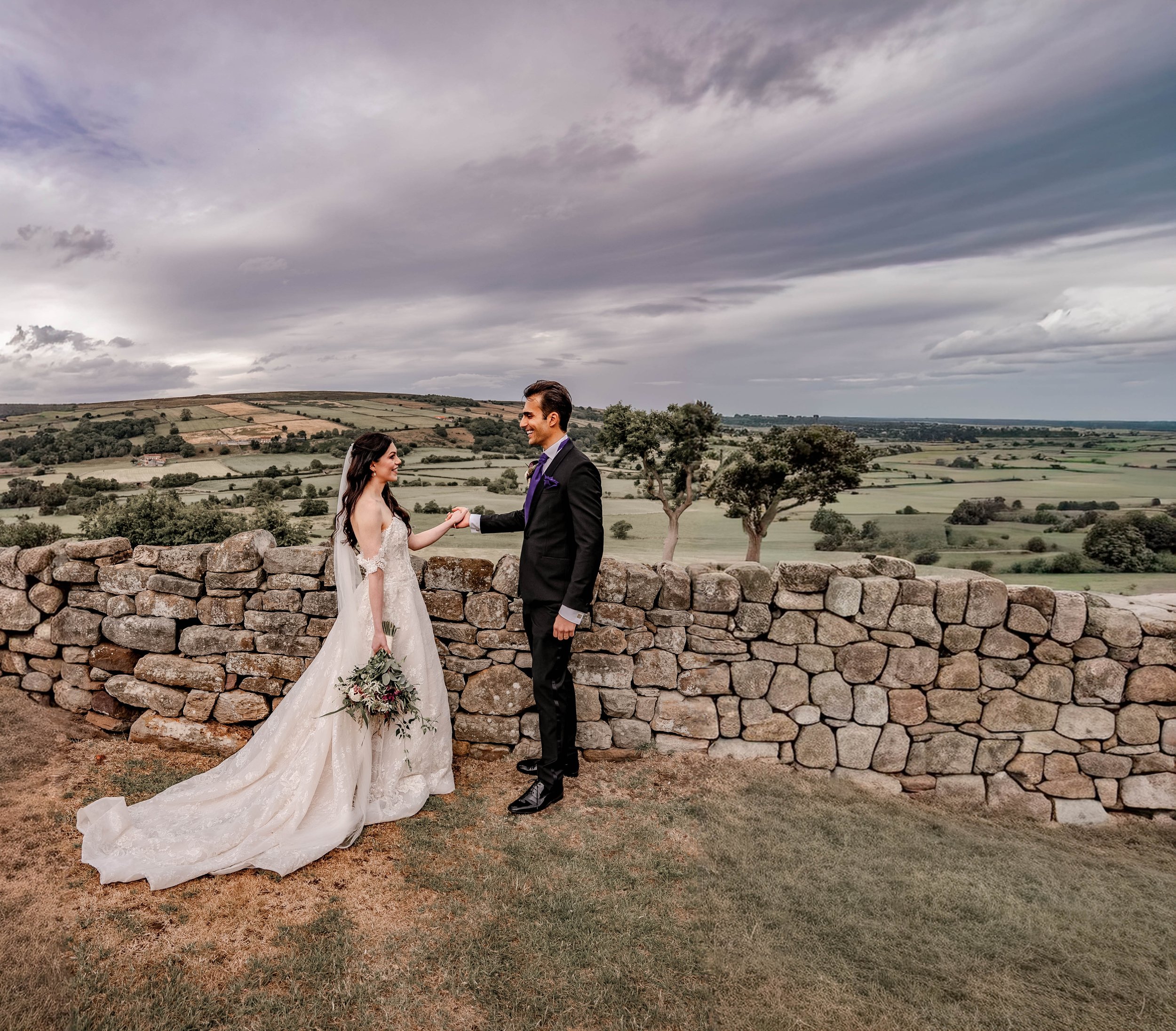 A bride and groom holding hands and smiling, standing outdoors in front of a stone wall with a landscape of rolling hills and cloudy sky in the background.