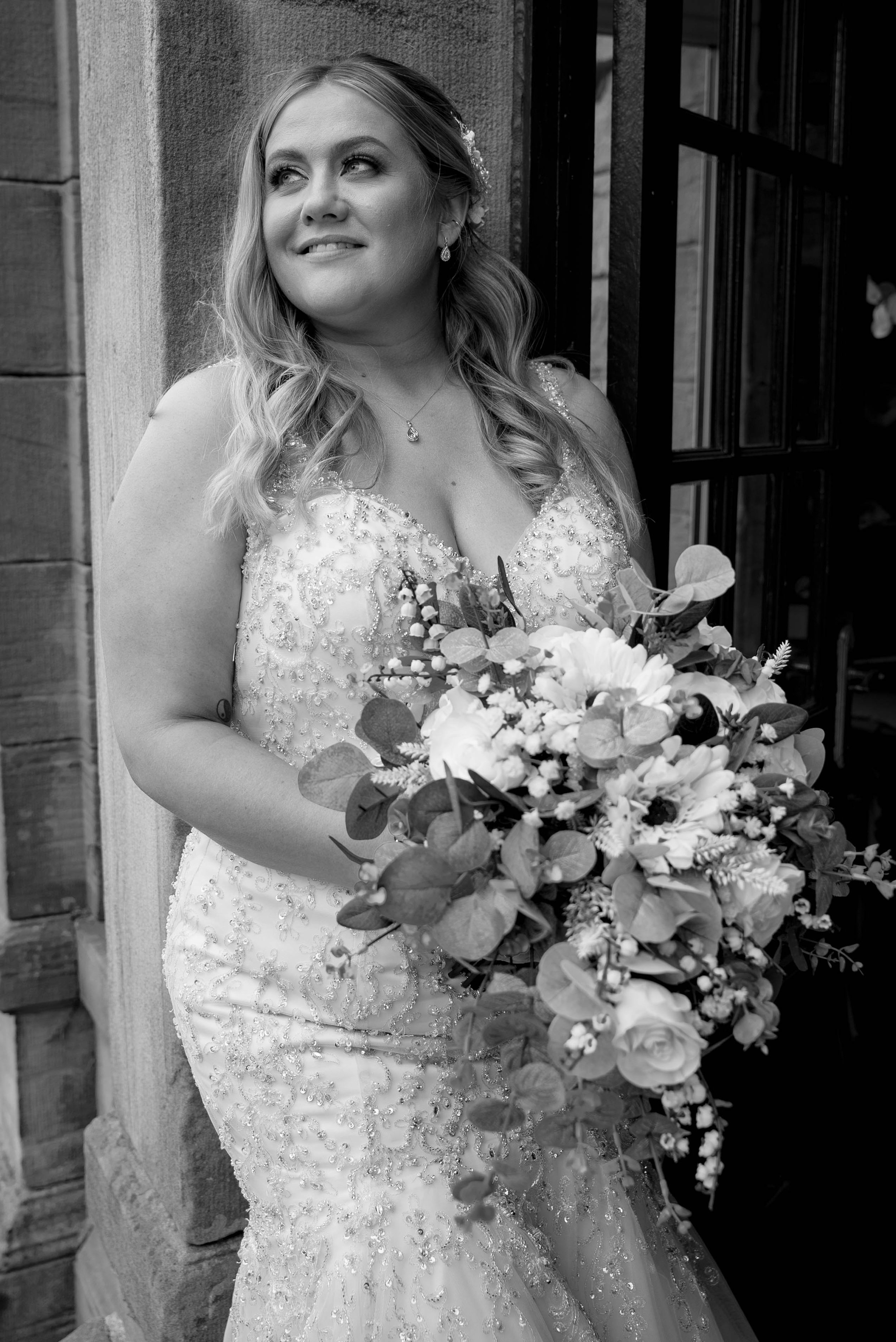 Black and white photo of a woman in a wedding dress holding a large bouquet of flowers, standing outdoors near a stone wall and window.