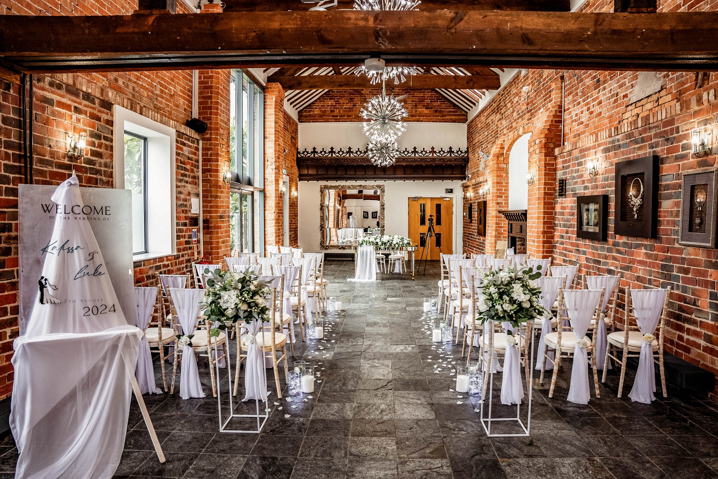 Indoor wedding ceremony setup with rows of white-draped chairs and floral arrangements, candles, and a welcome sign on an easel, in a brick-walled venue with large windows and decorative lighting fixtures.