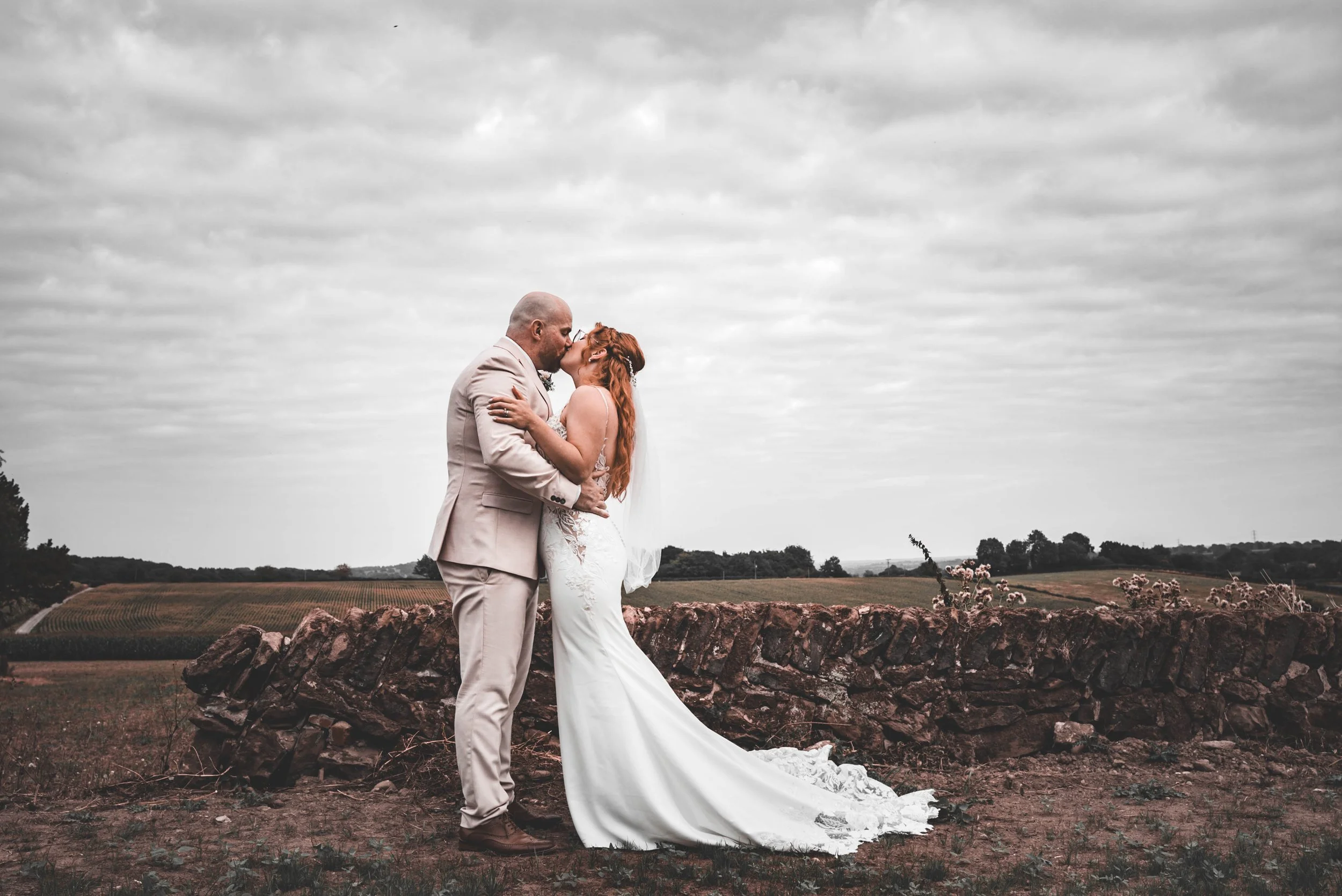 A couple in wedding attire sharing a kiss outdoors, with a stone wall and rolling hills in the background under a cloudy sky.