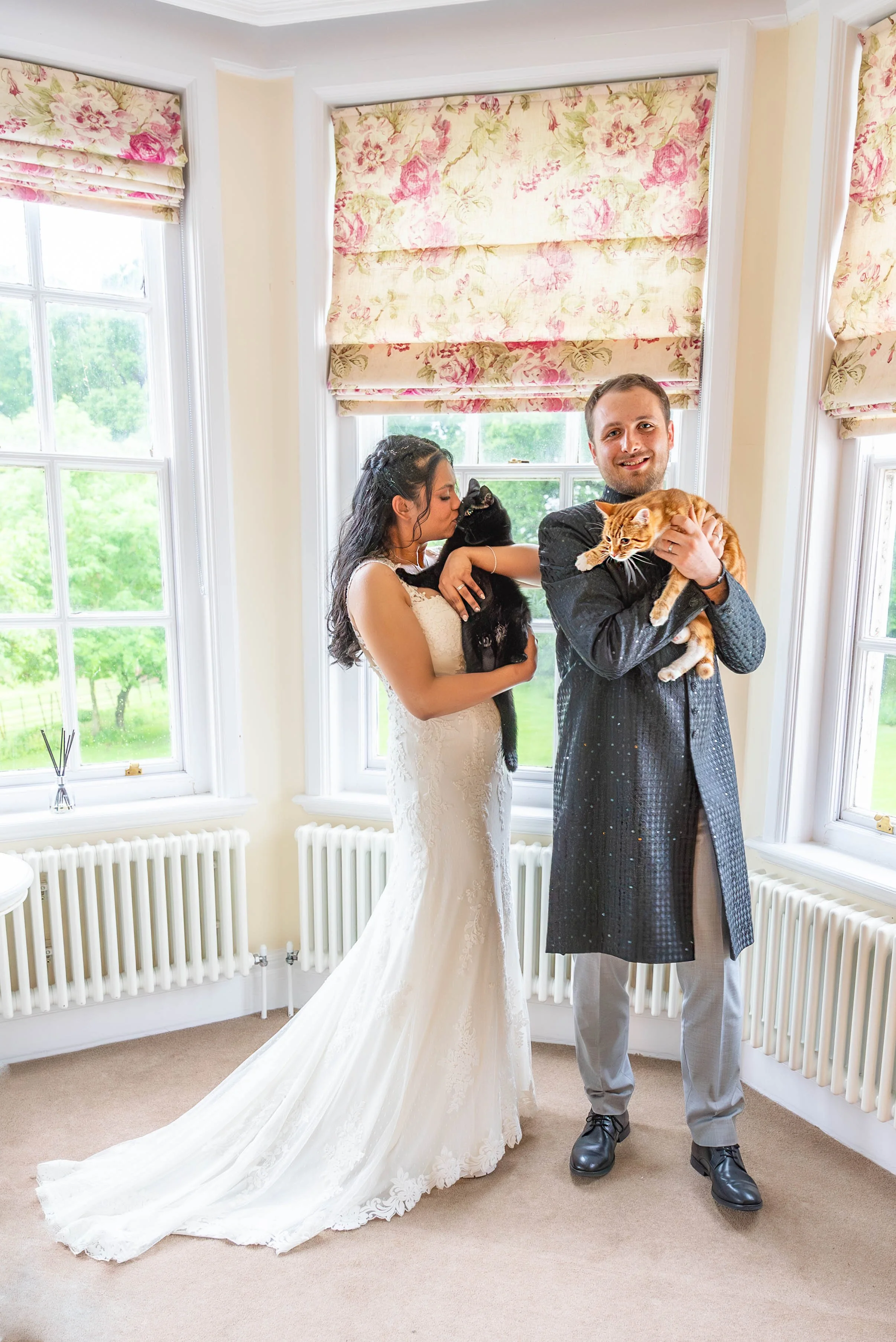Bride and groom holding cats indoors near large windows with floral curtains.